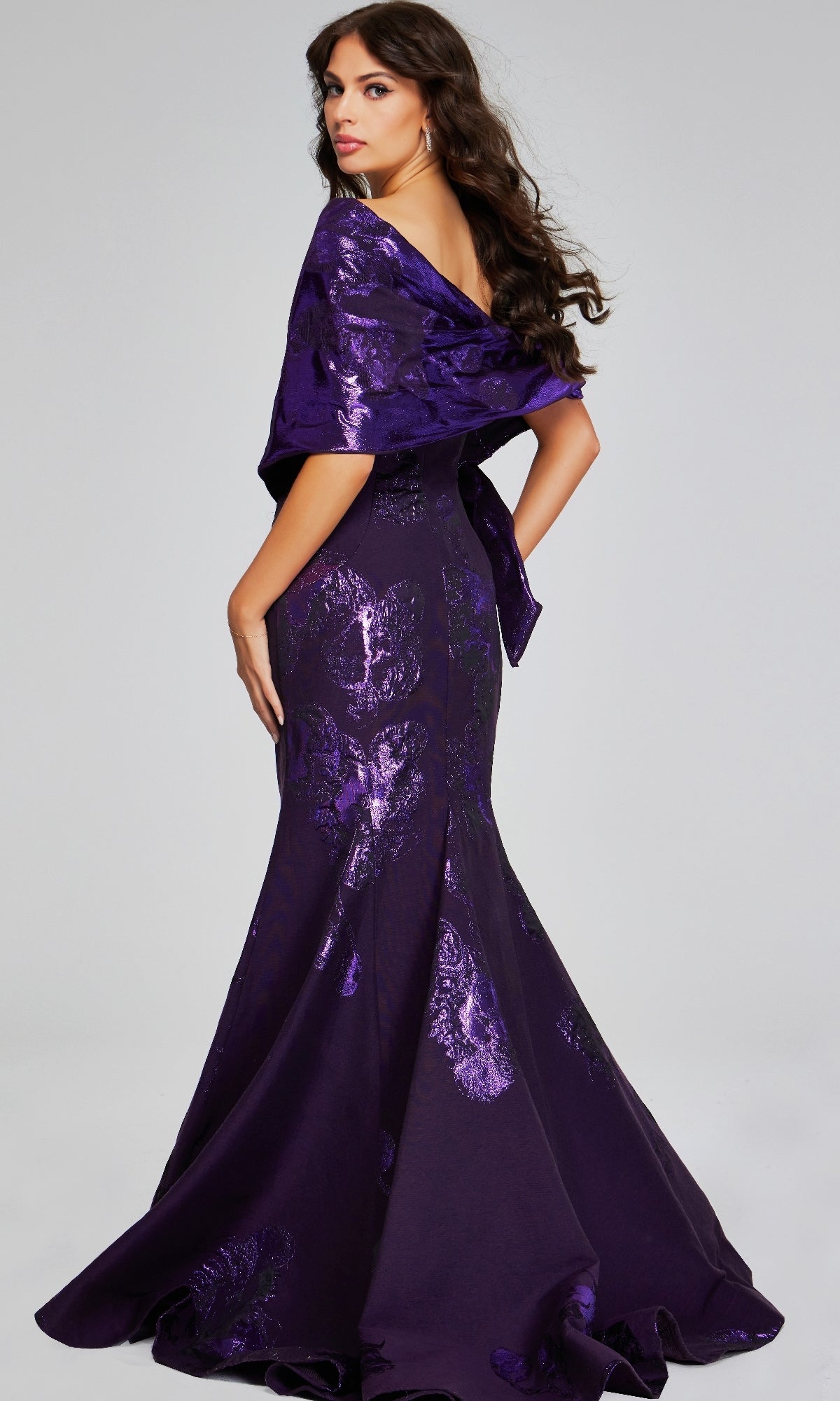 A woman stands looking over her shoulder in a floor-length, off-the-shoulder purple Jovani 40318 long formal dress with shimmering patterns.