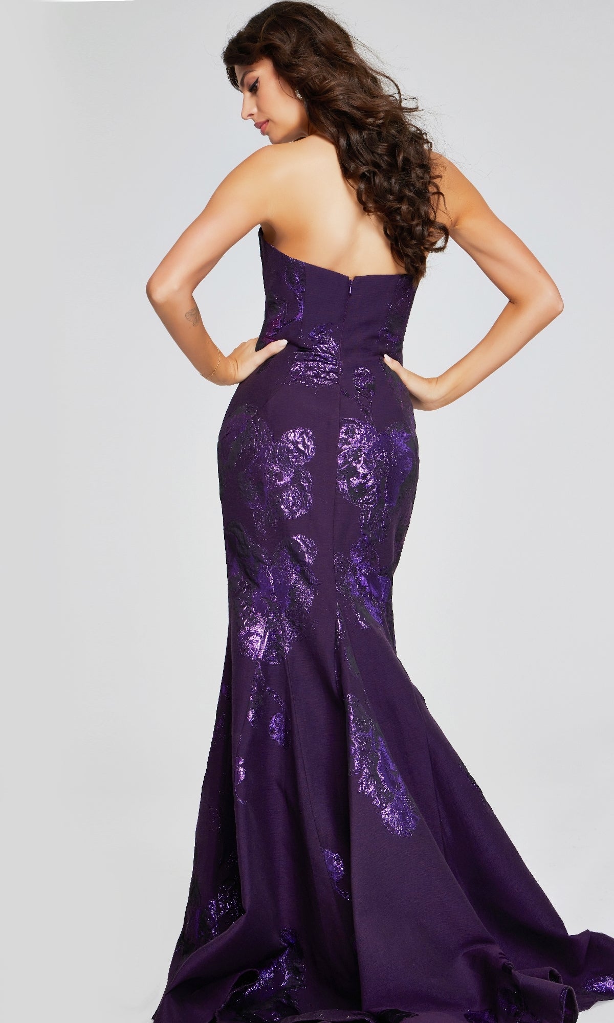 Woman in a strapless, floor-length Jovani 40318 mermaid gown with shiny floral patterns stands with her back to the camera, hands on hips.