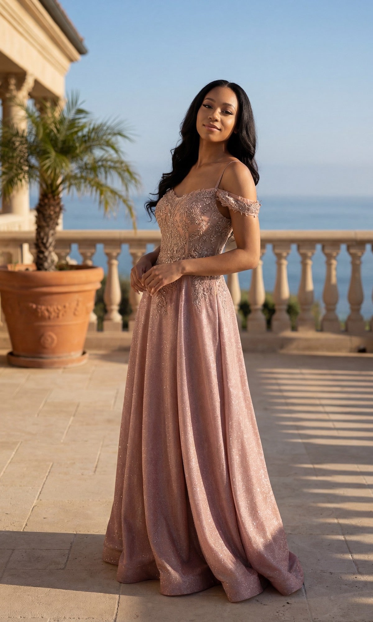 A woman in the Long Prom Dress: Chicas 4031, a floor-length off-shoulder rose gown with an embroidered bodice, stands on a sunlit terrace with a potted palm, balustrade, and ocean view in the background.