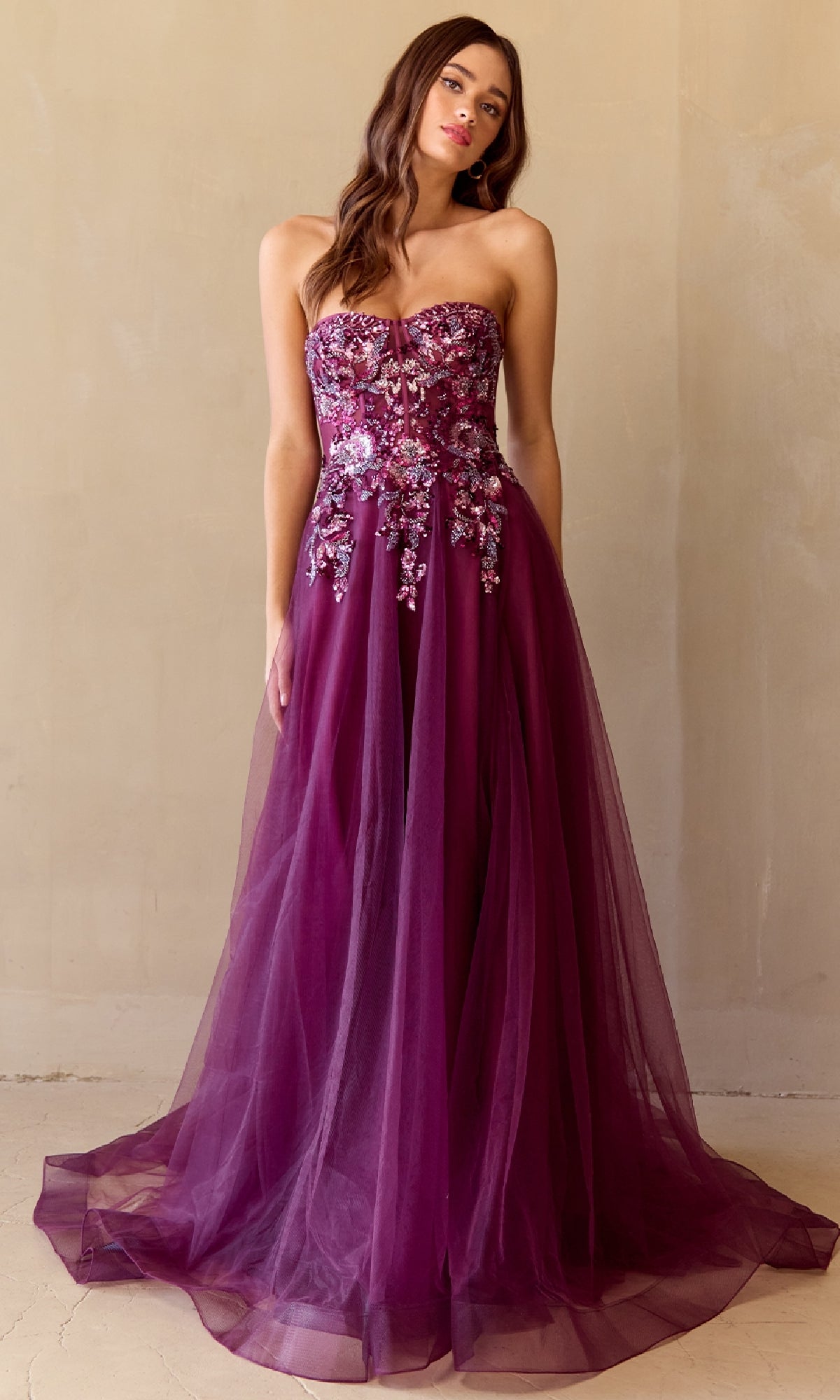 A woman wears the Amelia Couture 4031 Strapless A-Line Prom Gown with floral appliqués on the bodice and a tulle skirt, standing against a plain beige wall.