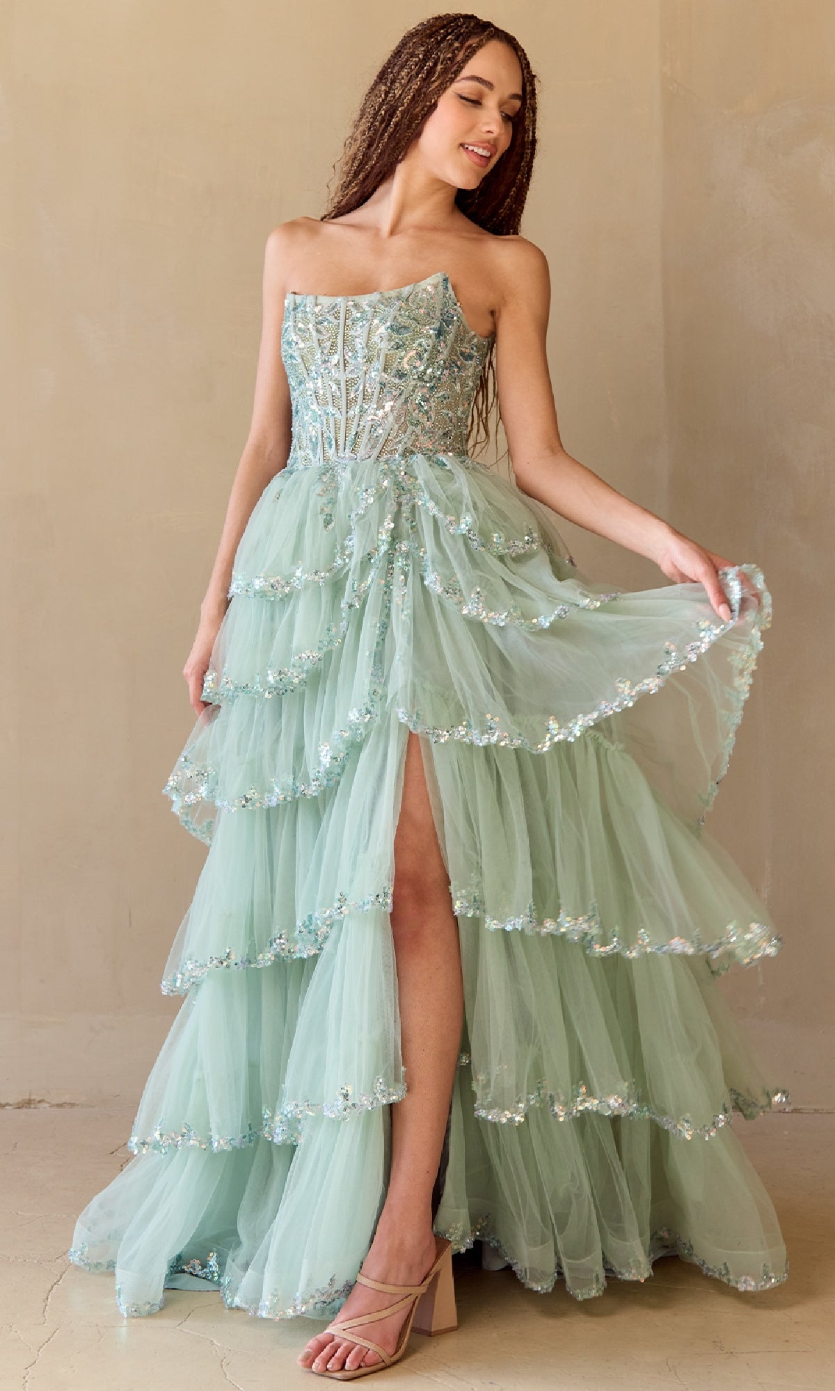A woman stands indoors, holding one side of her mint green tulle Ruffled Strapless Prom Dress: Amelia Couture 4033, featuring a sequin corset bodice, dramatic ruffles, and a high slit.