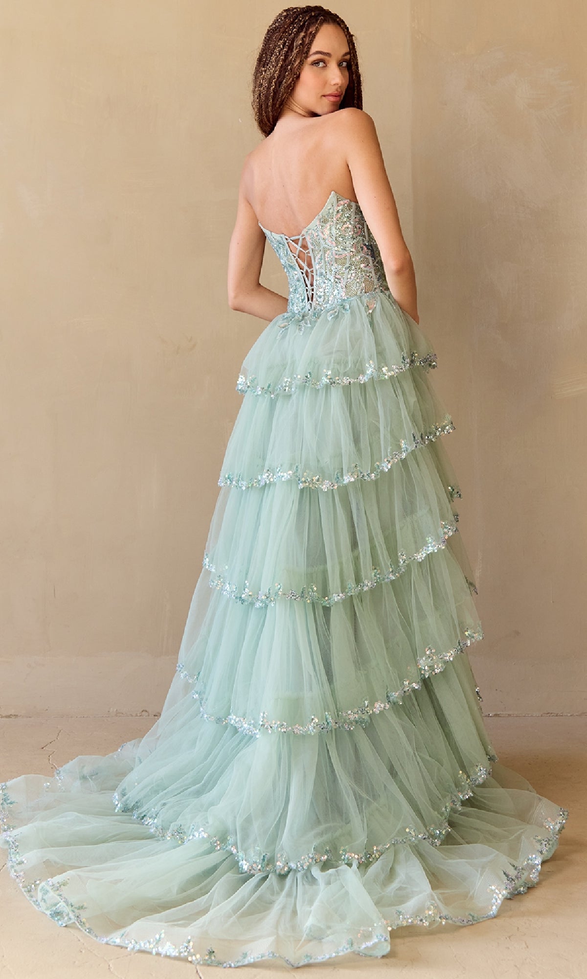 A woman models the Amelia Couture 4033 Ruffled Strapless Prom Dress in light green, featuring floral embellishments, a sequin corset bodice, and a tiered tulle skirt, posed against a plain beige background.