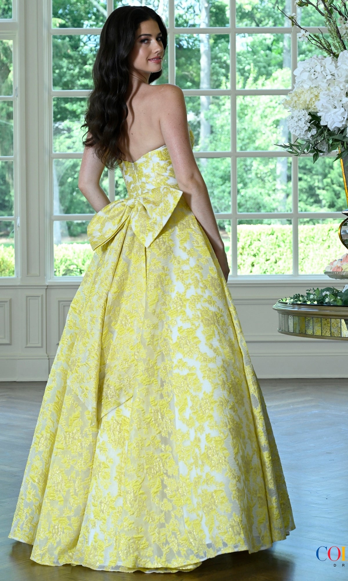 A woman wears a Colors Dress 4039, a floor-length, strapless yellow floral jacquard prom dress featuring a large bow at the back.
