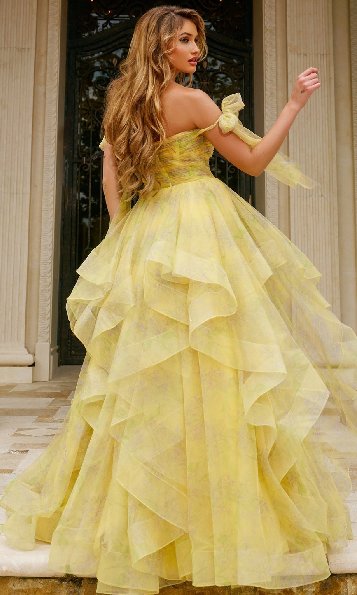 A woman with long wavy hair poses in front of a columned building wearing the Jovani 40400 long prom dress, featuring off-the-shoulder straps, a printed design, and a layered ball-gown skirt.