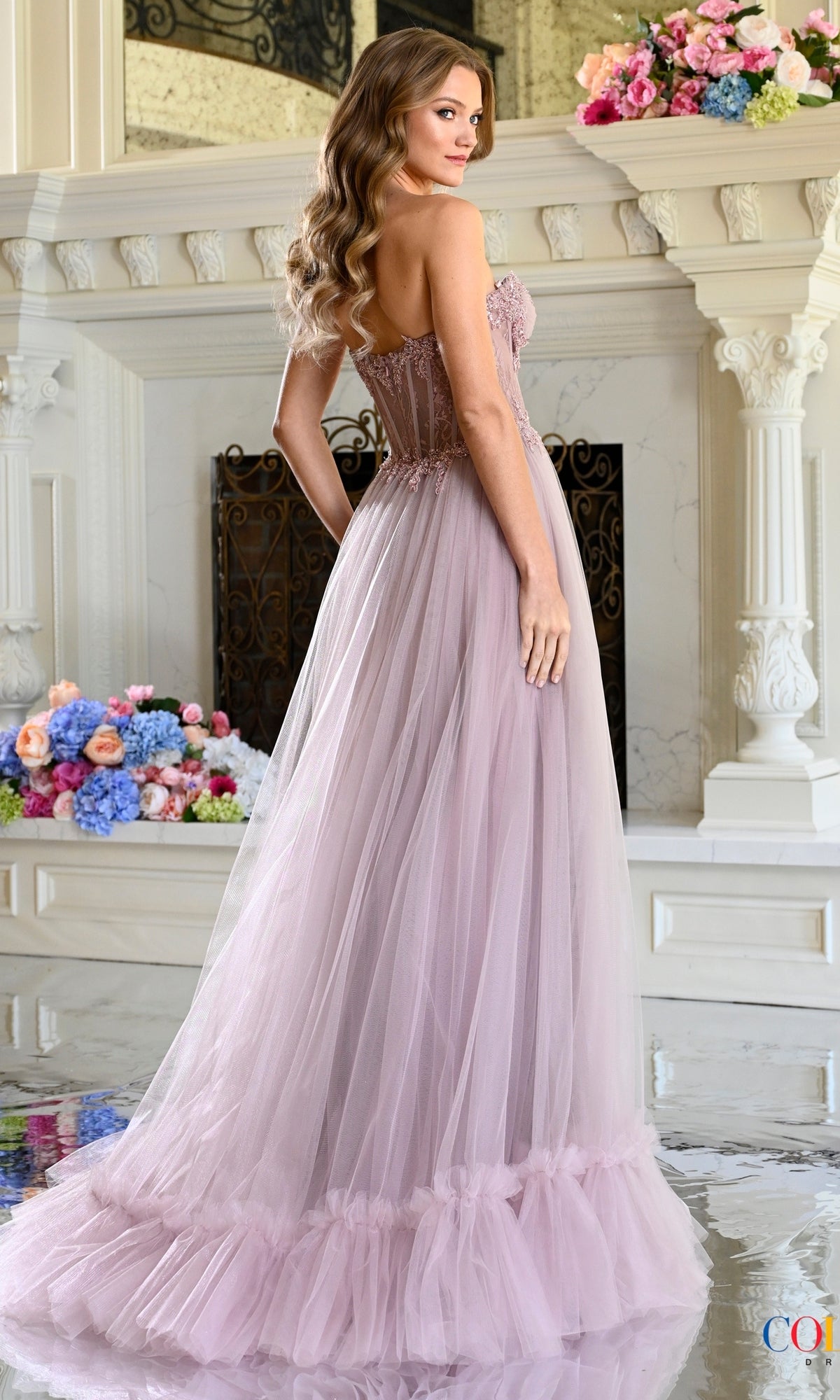 A woman wearing a Long Prom Dress: Colors Dress 4041, featuring a floor-length lavender design with a sheer lace corset bodice and a ruffled tulle skirt, standing before a floral-decorated fireplace.