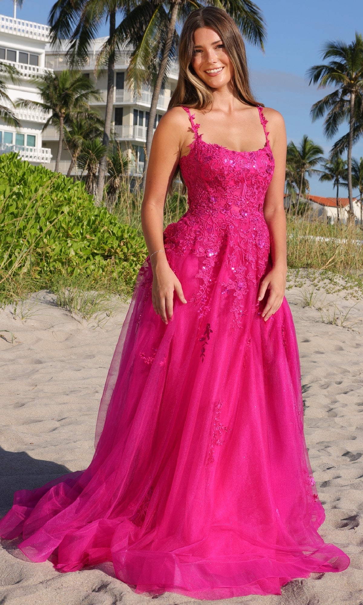 A woman wears the Amelia Couture 4044 Long Corset Prom Ball Gown, a bright pink, floor-length A-line dress with floral embellishments, standing on a sandy beach with palm trees and buildings in the background.