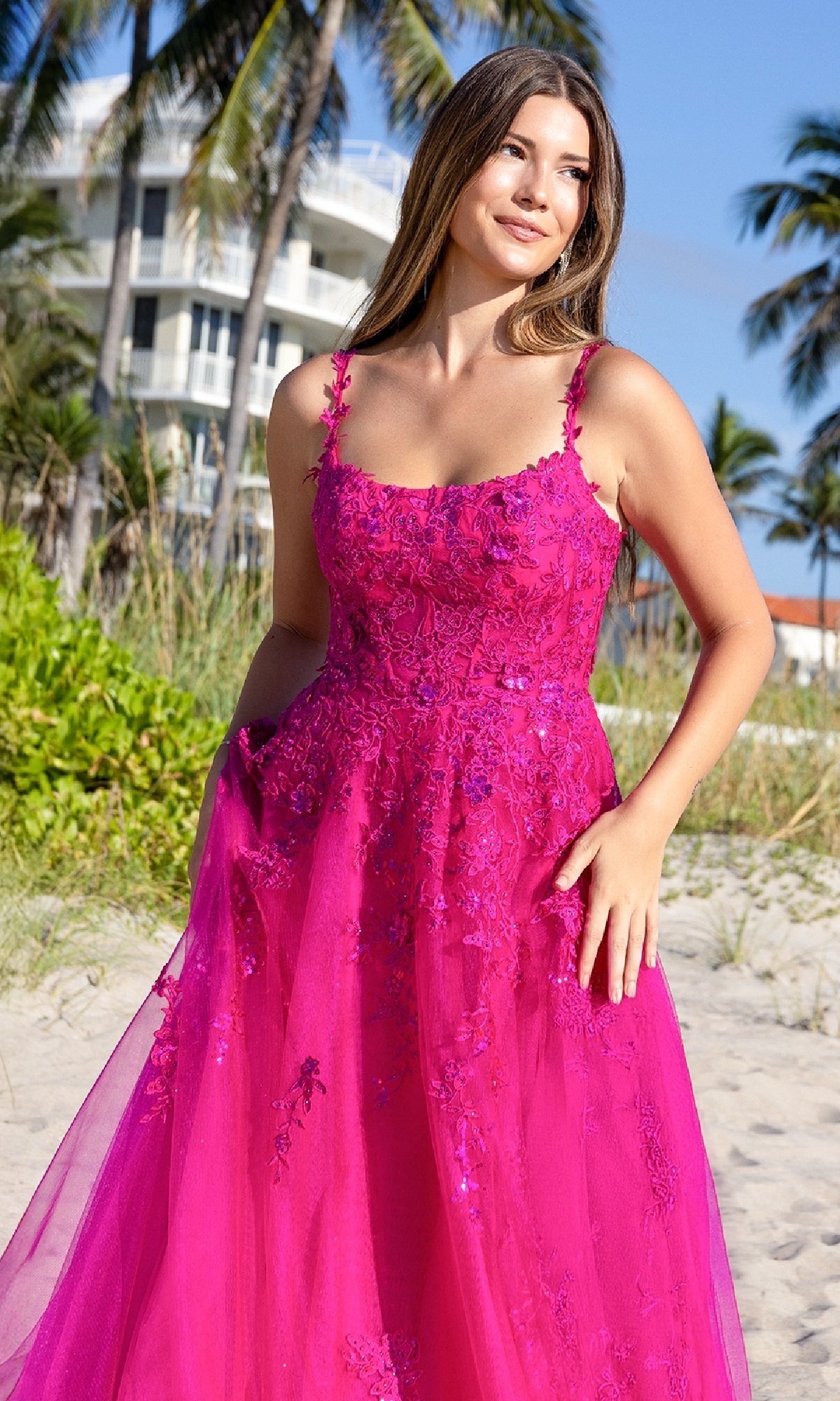 A woman in the Amelia Couture 4044 Long Corset Prom Ball Gown stands on a sandy beach with palm trees and buildings in the background.