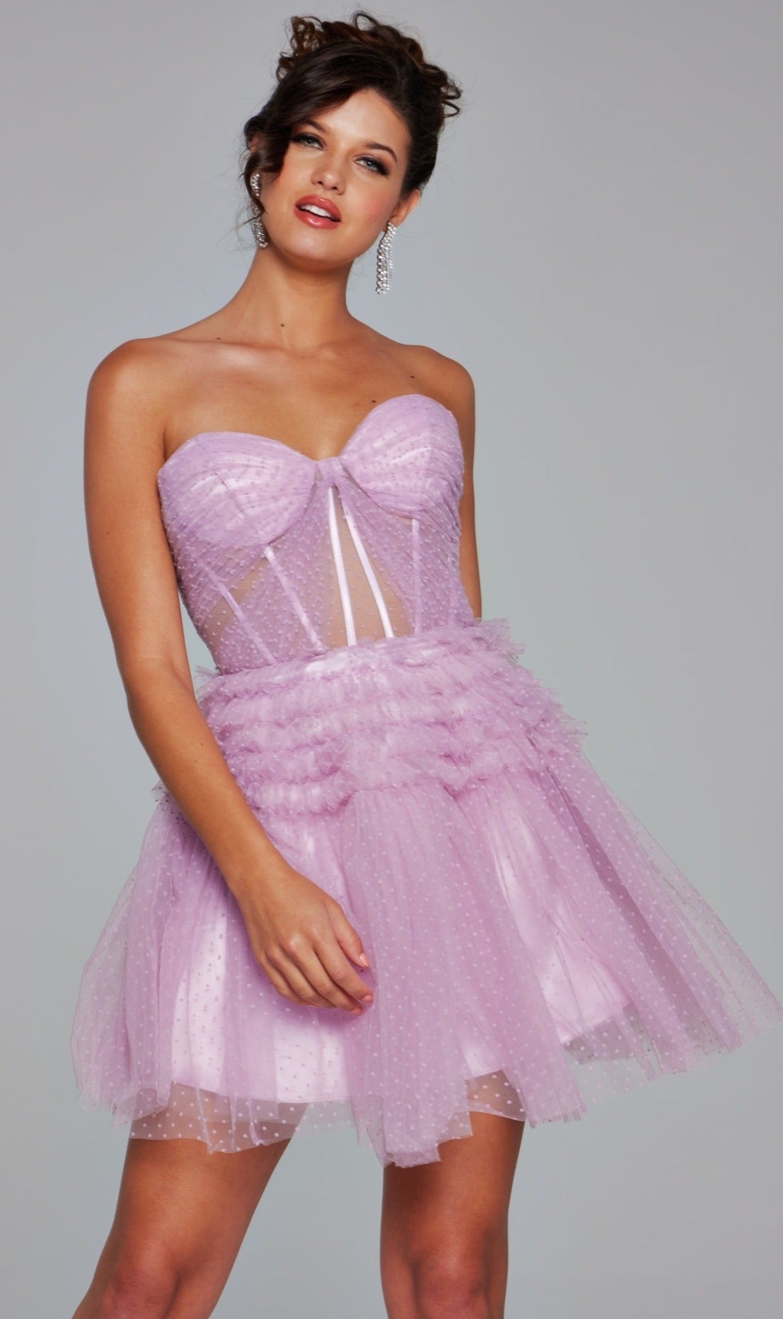 A woman stands against a plain background wearing the Short Homecoming Dress 40467 by Jovani, a strapless light purple tulle mini dress with a fitted sheer corset bodice and layered skirt.