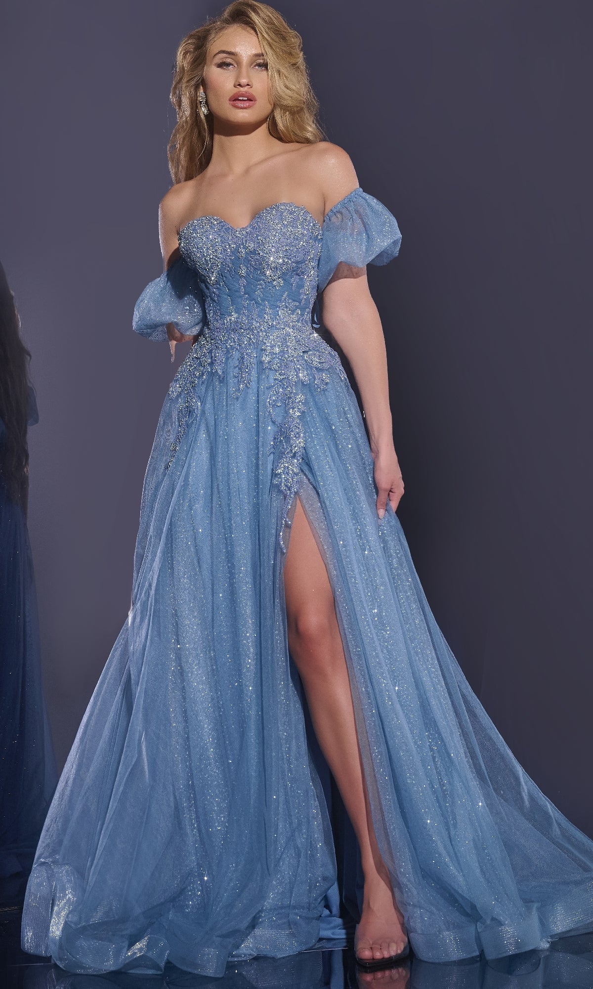 A woman stands against a dark background, wearing the Jovani 40474 long blue prom dress with puff sleeves, floral embellishments, and a high leg slit.