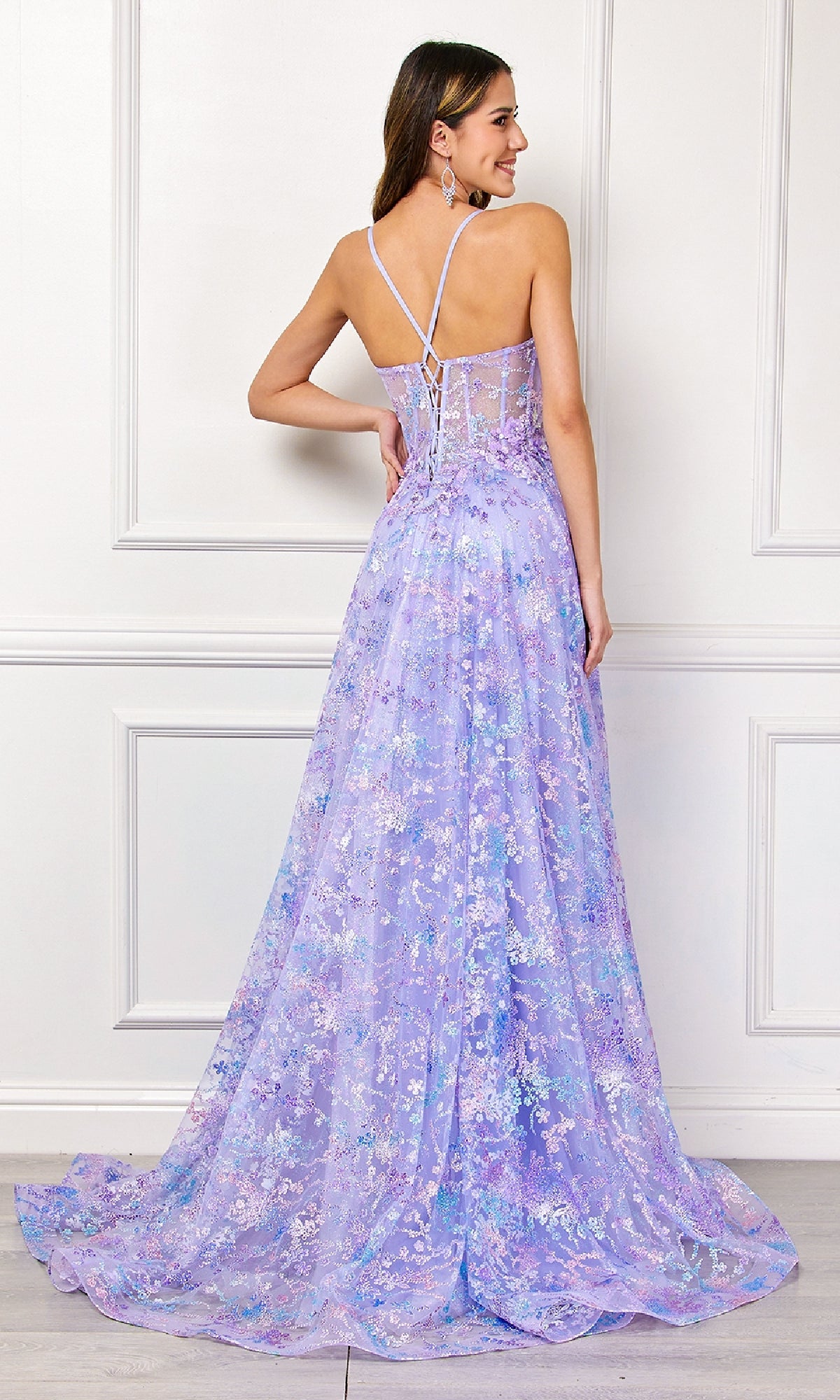 Woman wearing the Embellished Long Prom Dress: Amelia Couture 4050, an elegant floor-length lavender gown with floral design, sheer lace-up back, and spaghetti straps, standing before a white paneled wall.