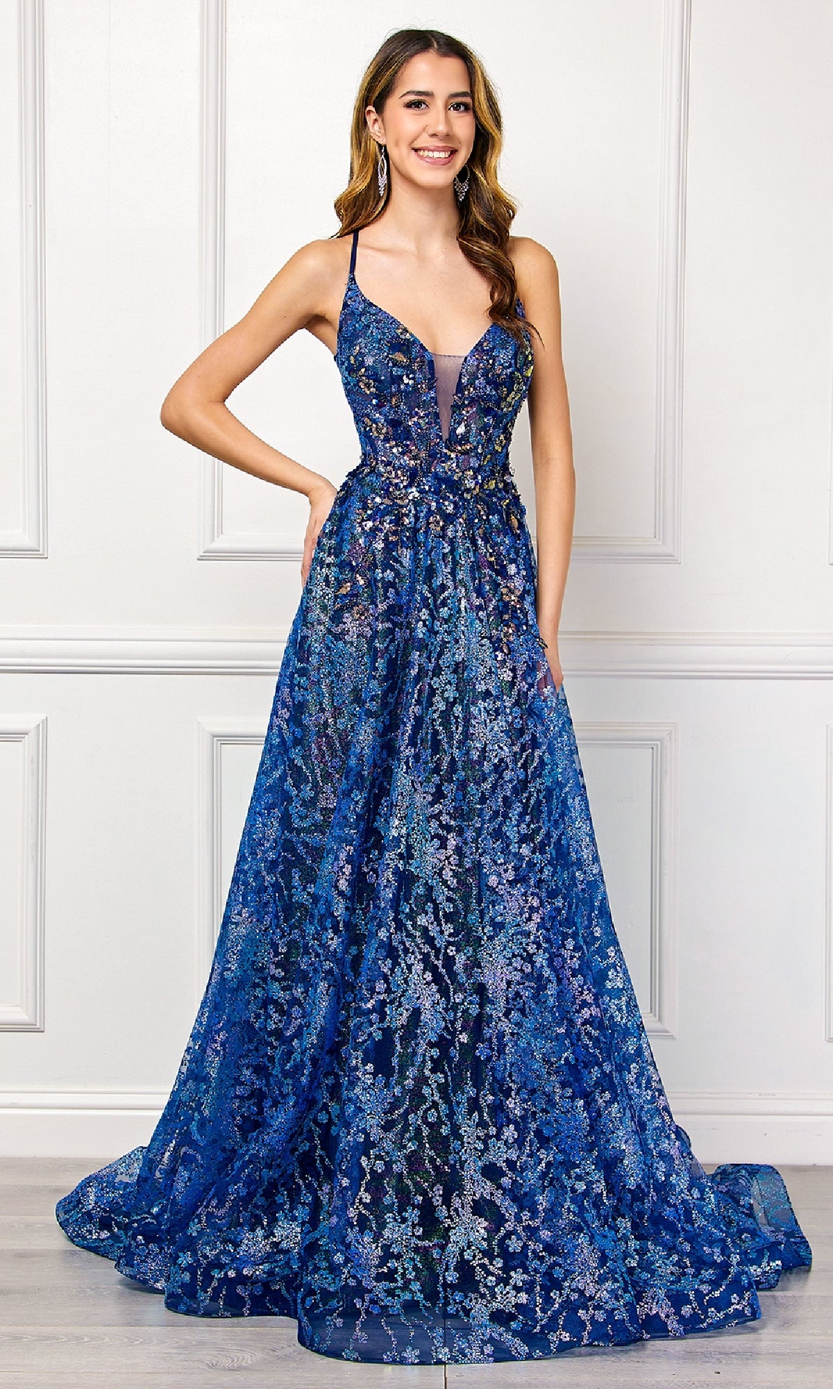 A woman stands before a white paneled wall in the Amelia Couture 4050—a floor-length, sleeveless embellished long prom dress in deep blue with floral patterns and a plunging neckline.