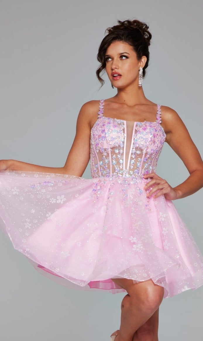 A woman models the Short Homecoming Dress 40501 by Jovani, a light pink A-line dress with floral sequin embroidery, sheer accents, and spaghetti straps, holding out the skirt against a plain gray background.