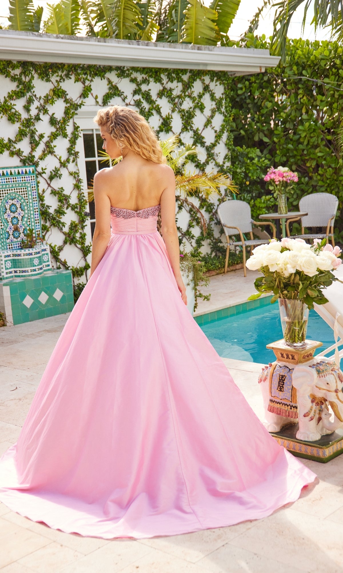 A woman in the Ava Presley 40523 long strapless prom dress stands by a pool, facing away, with greenery, flowers, and patio furniture in the background.
