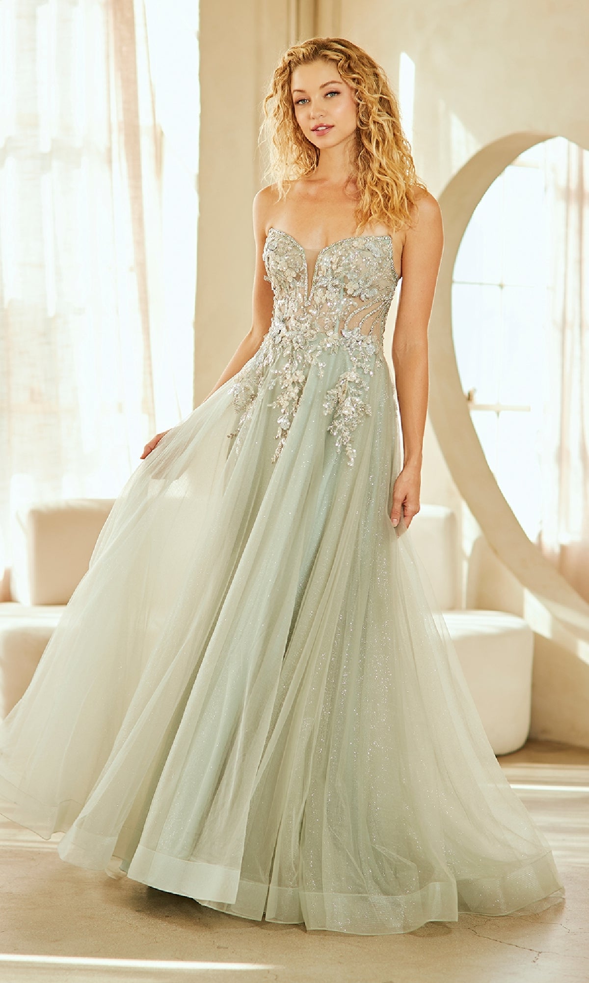A woman stands indoors in natural light wearing the Strapless Glitter Prom Gown: Amelia Couture 4062, featuring a strapless design, glitter details, and 3-D floral appliques.