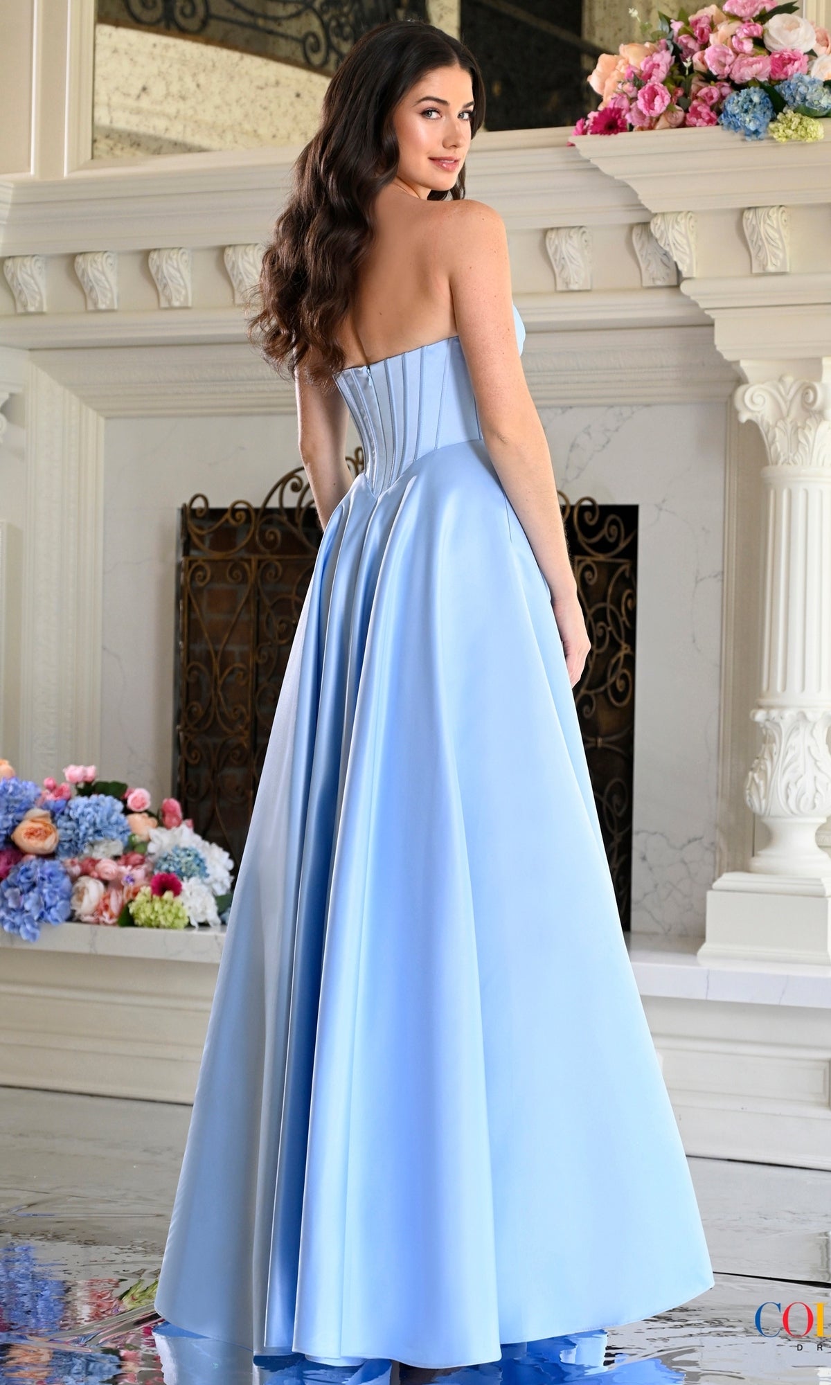 A woman wearing a light blue Colors Dress 4063 long prom dress, featuring a strapless, floor-length gown with a pleated sweetheart neckline and a corset-style bodice.