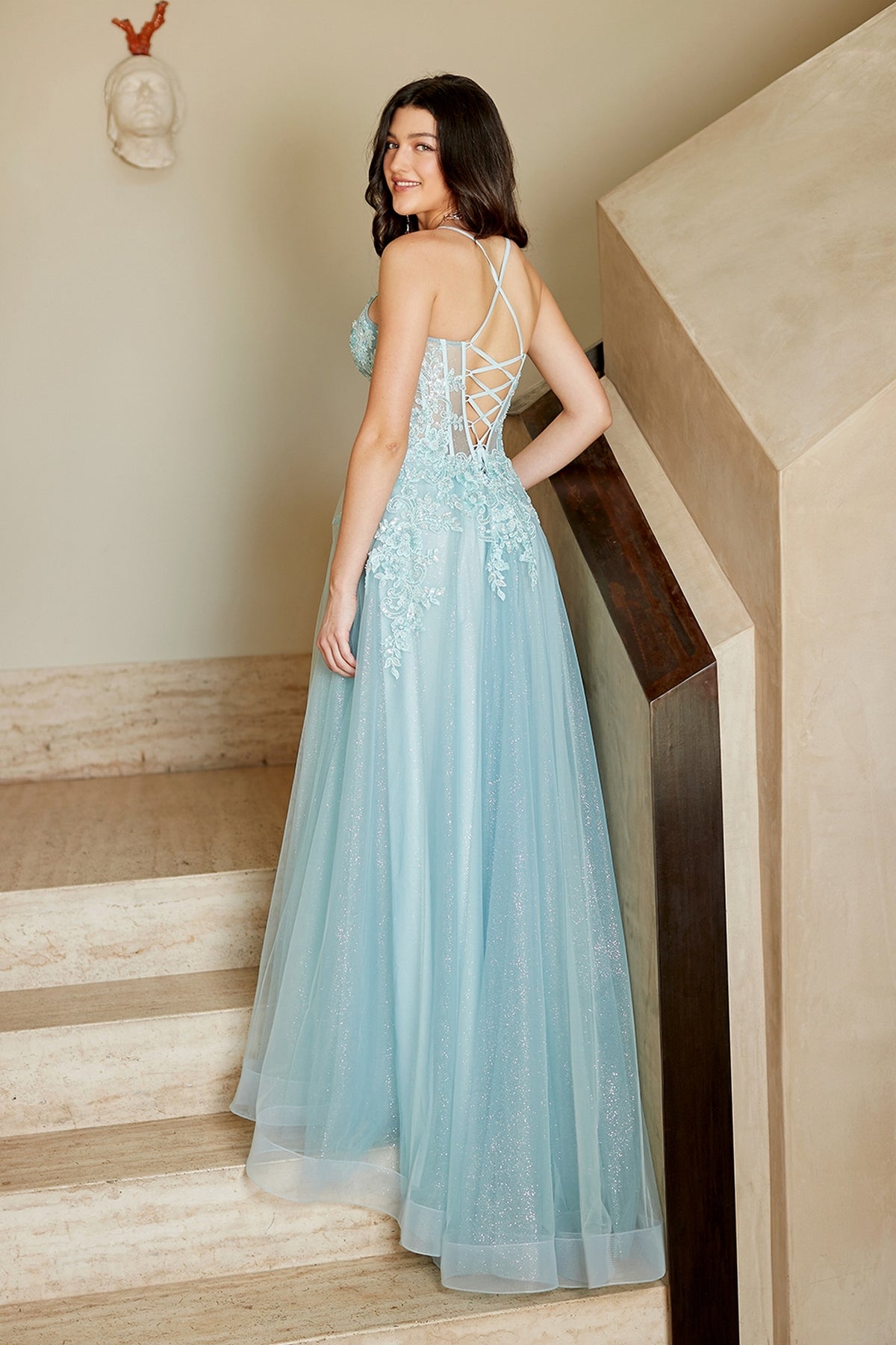 A person on a staircase wearing a light blue, floor-length lace appliqué Long Prom Dress: Amelia Couture 4074 with a lace-up back.