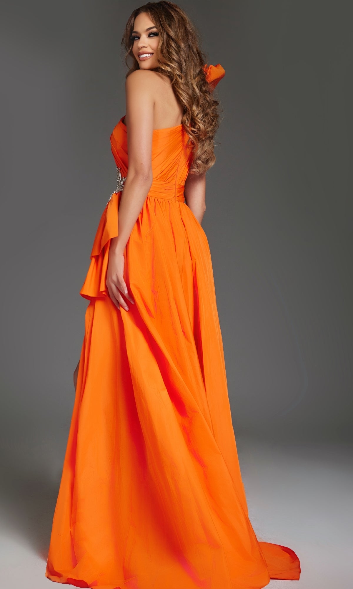 A woman wearing the Jovani 40929 long, strapless orange prom dress poses against a plain gray background, looking over her shoulder with a smile.