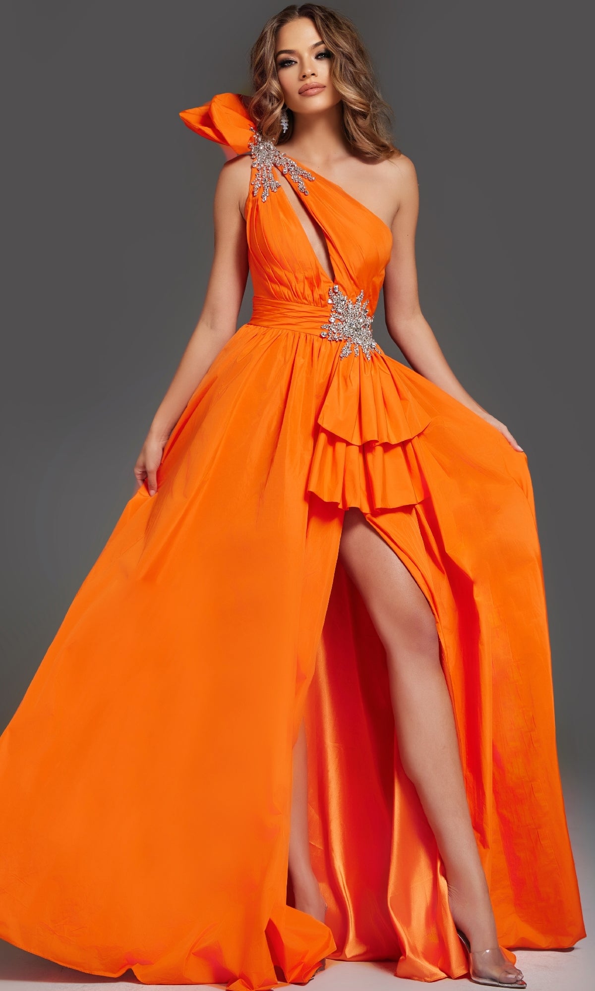A woman wearing the Jovani 40929 long prom dress, featuring a bright orange hue, one-shoulder neckline, ruffle accents, jeweled embellishments, and a high leg slit poses confidently.