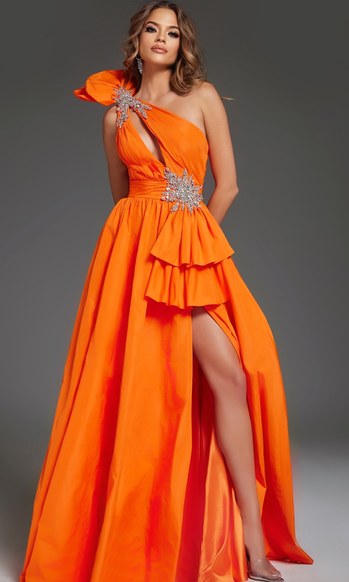 A woman stands against a gray background wearing the Jovani 40929 long prom dress, featuring bright orange color, a one-shoulder neckline, ruffles, jeweled embellishments, and a thigh-high slit.