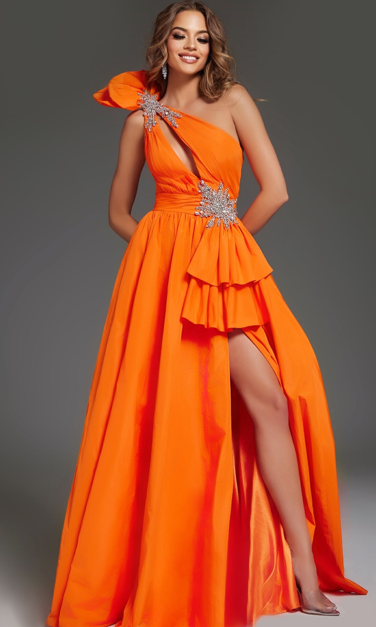 A woman models the Jovani 40929 long prom dress in bright orange, featuring a one-shoulder neckline, embellished details, and a thigh-high slit, standing against a plain gray background.