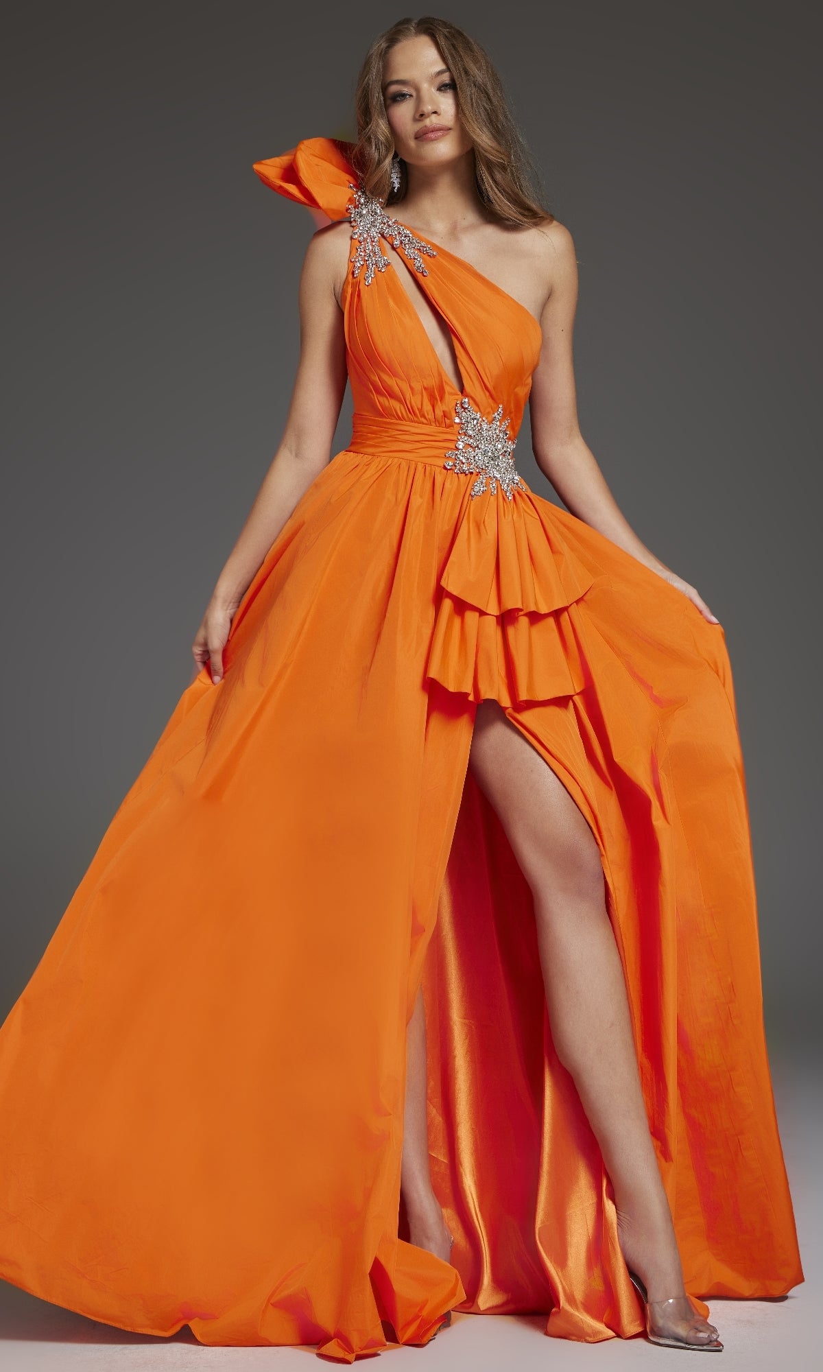 A woman poses against a plain gray background wearing the Jovani 40929—a bright orange, one-shoulder long prom dress with silver embellishments and a high slit.