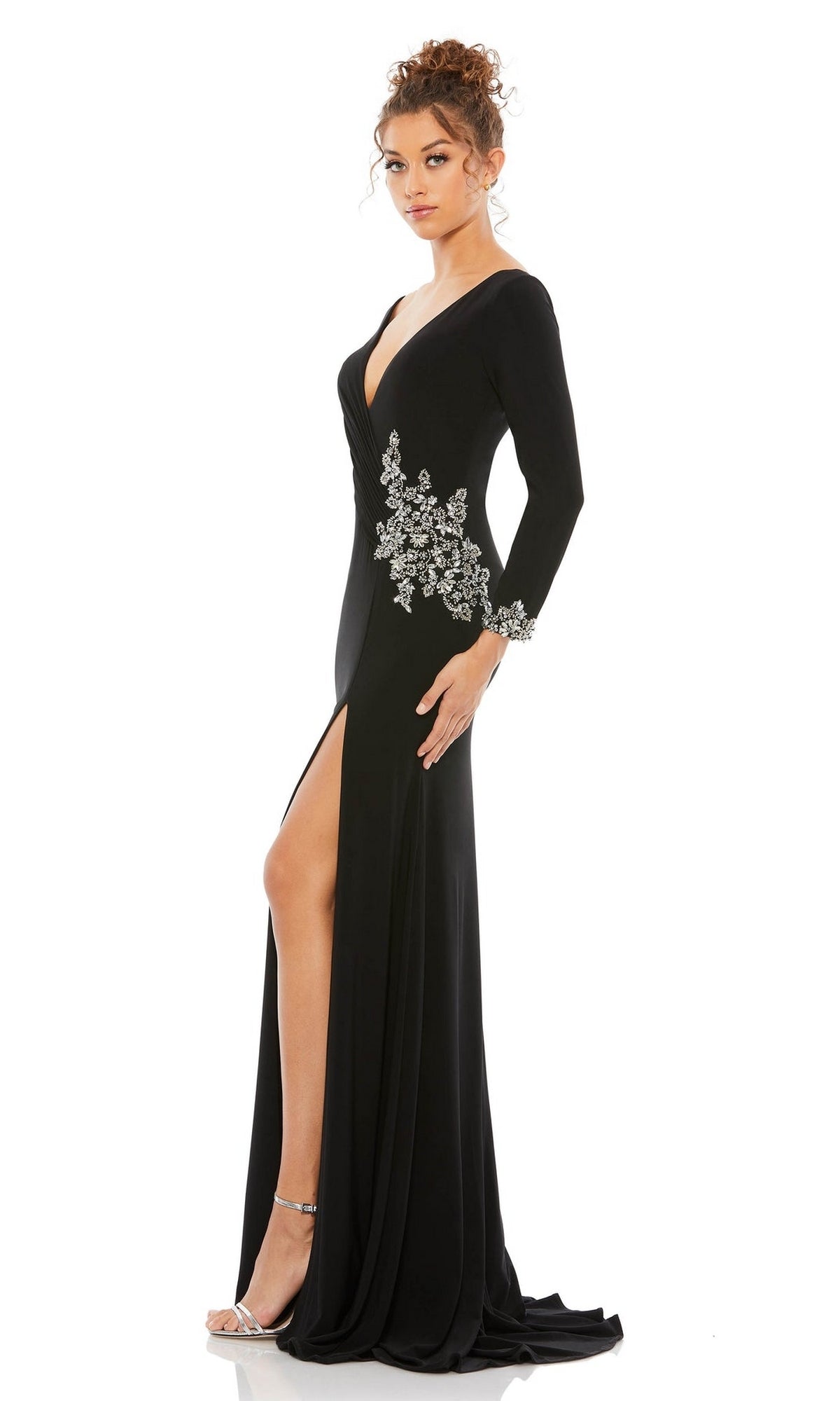 A woman models the Long Formal Dress 41016 by Mac Duggal—a black gown with long sleeves, a thigh-high slit, and silver floral embellishments—against a white background.