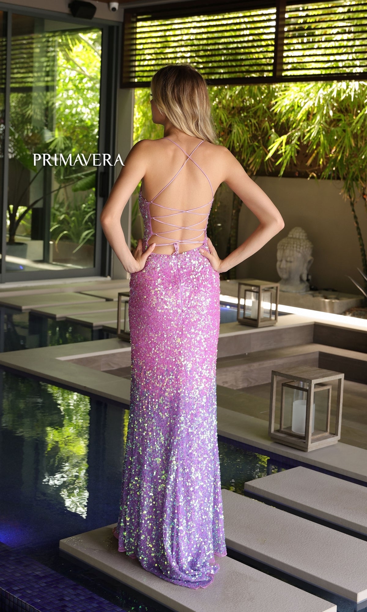 A woman stands beside an indoor pool wearing the Primavera Ombre Long Beaded Prom Dress 4102 in glittery pink and purple with crisscross straps, surrounded by plants, lanterns, and a Buddha statue.
