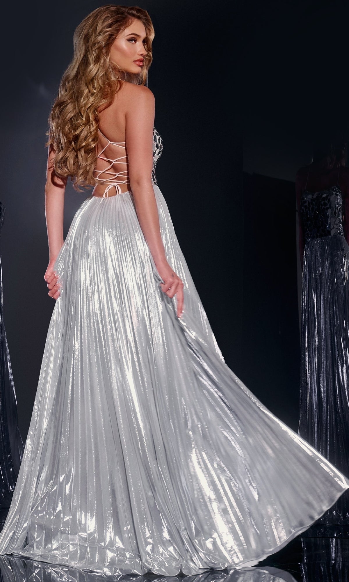 A woman with long wavy hair models the Jovani 41117 Long Prom Dress, a silver metallic gown with sequin embellishments and a lace-up back, looking over her shoulder against a dark background.