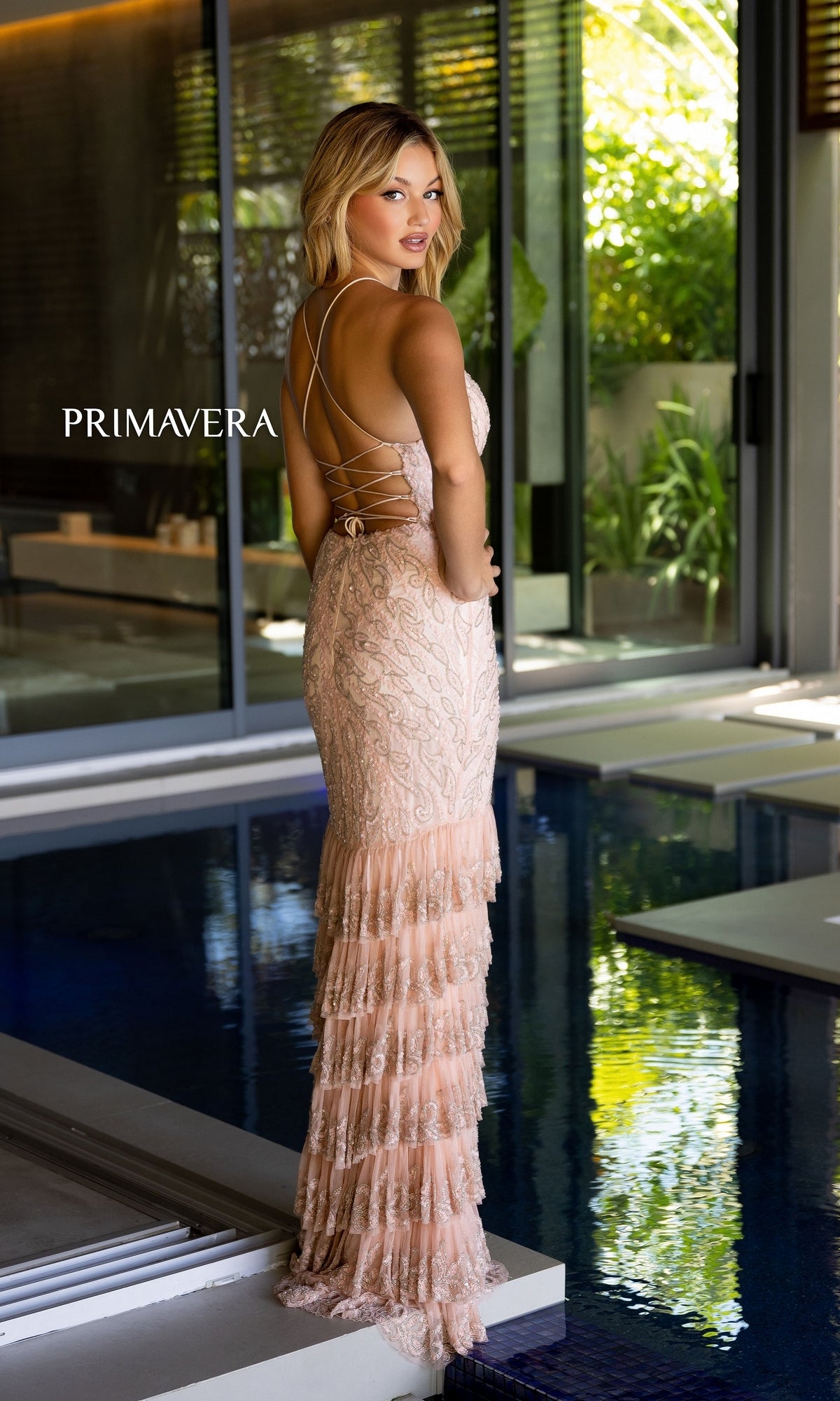 A woman in the Primavera Long Beaded Ruffled Prom Dress 4122, featuring light pink lace and tiered ruffles, stands by an indoor pool looking over her shoulder.