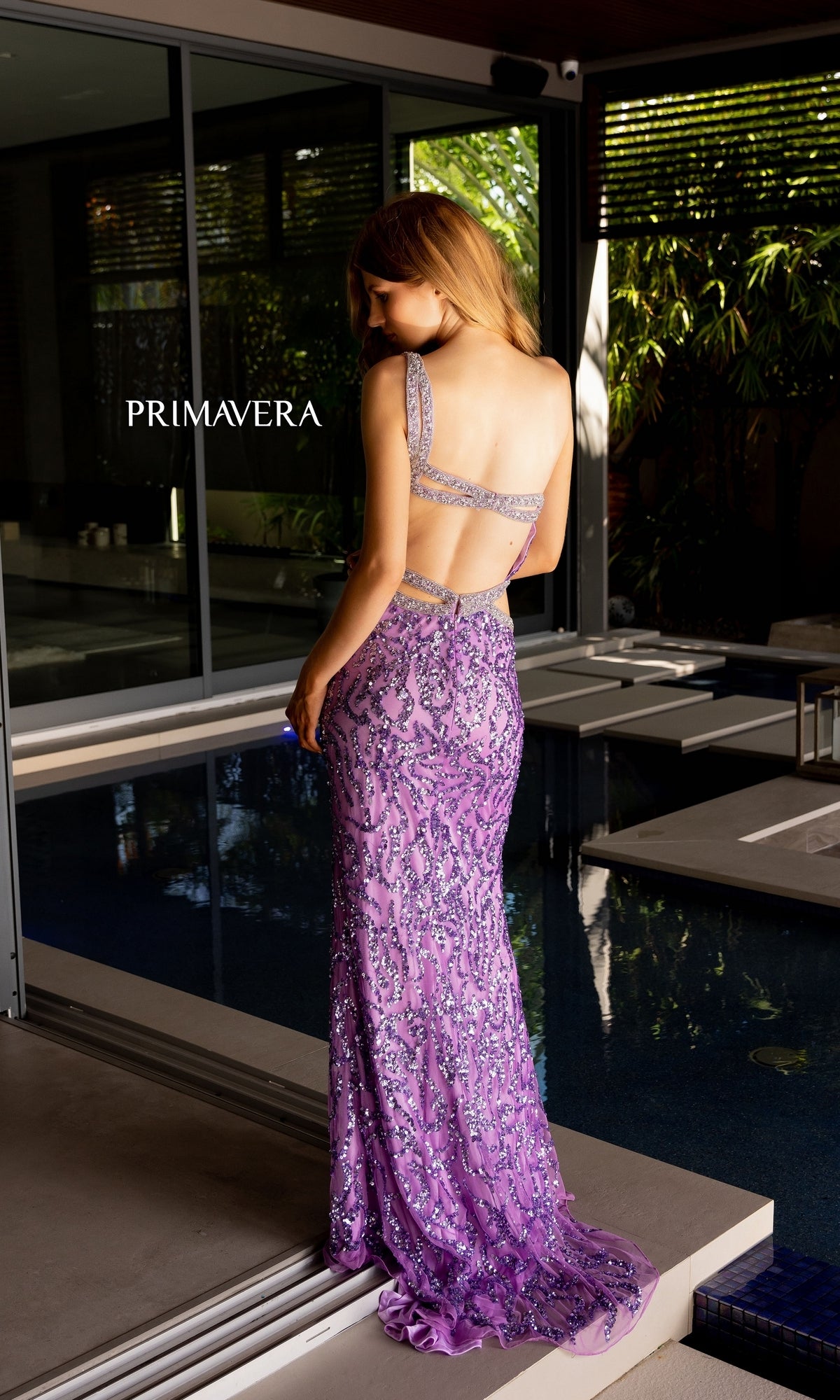 A woman stands by a pool wearing the Primavera One-Shoulder Beaded Long Prom Dress 4126. The floor-length, sleeveless lavender gown features sparkling beadwork, and