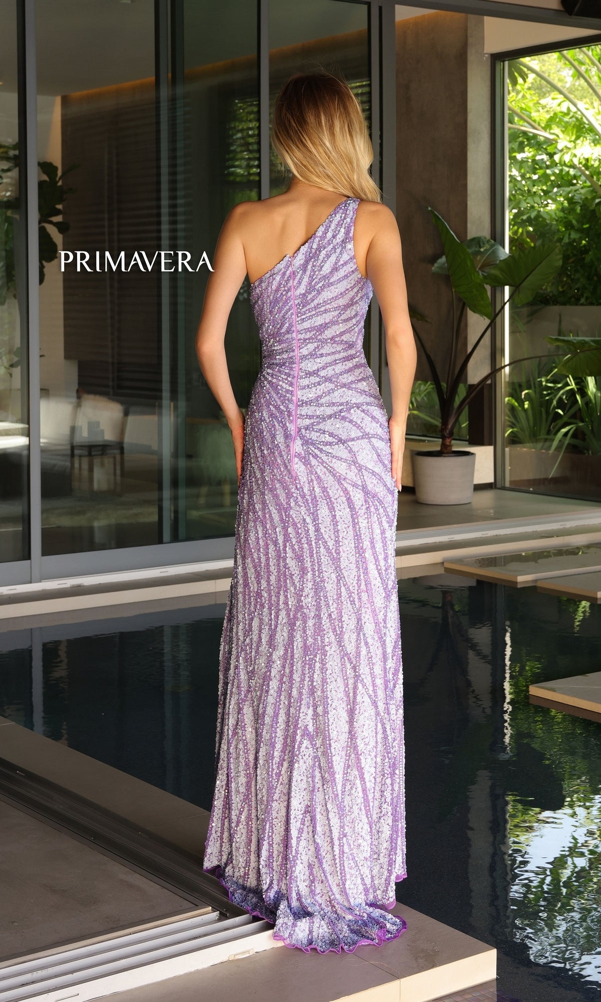 A woman stands indoors by a pool, wearing the Primavera One-Shoulder Long Beaded Prom Dress 4133 in shimmering lavender and silver. The photo, taken from behind, shows