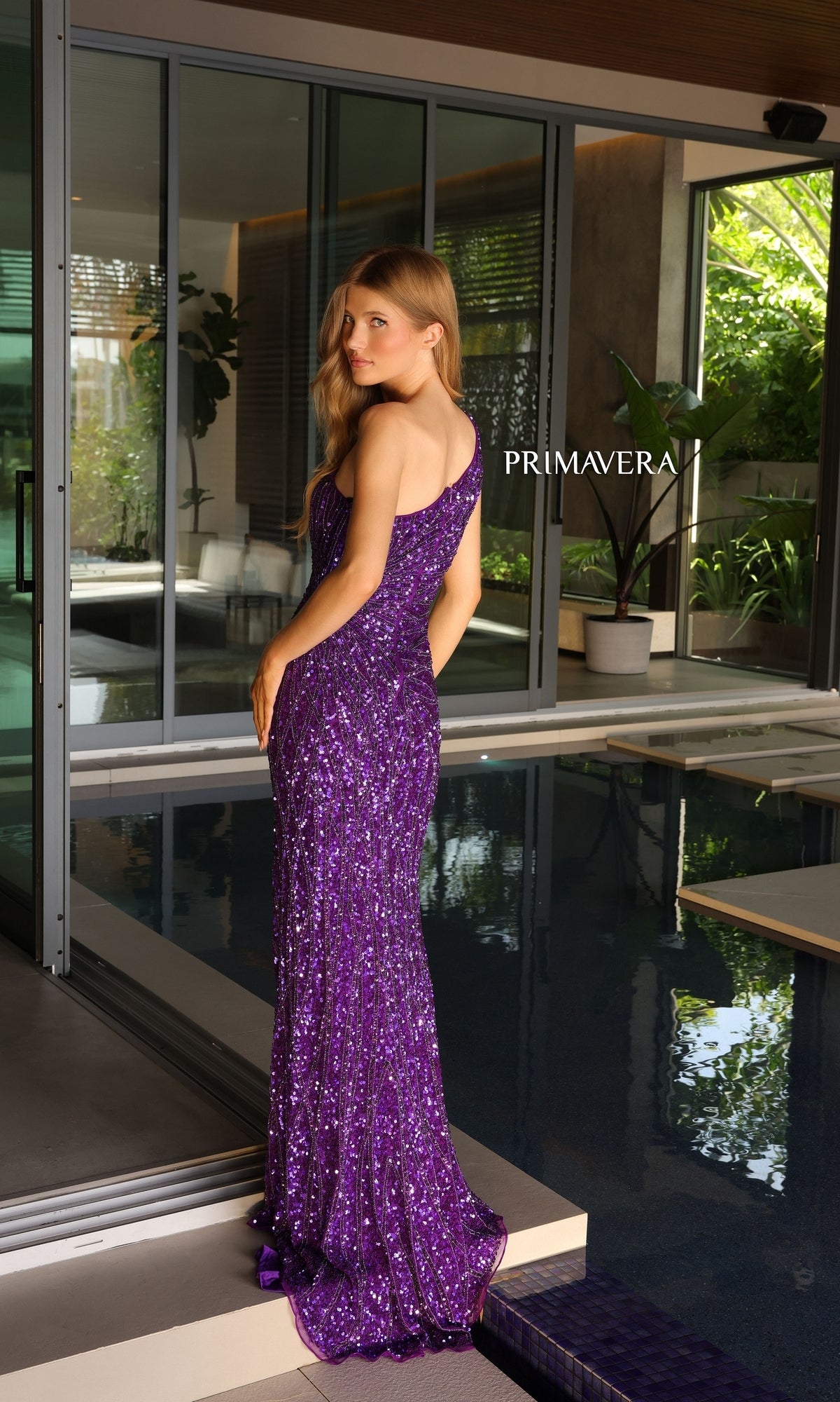 A woman in the Primavera One-Shoulder Long Beaded Prom Dress 4133 stands by an indoor pool, facing slightly away, with greenery and modern glass doors in the background.