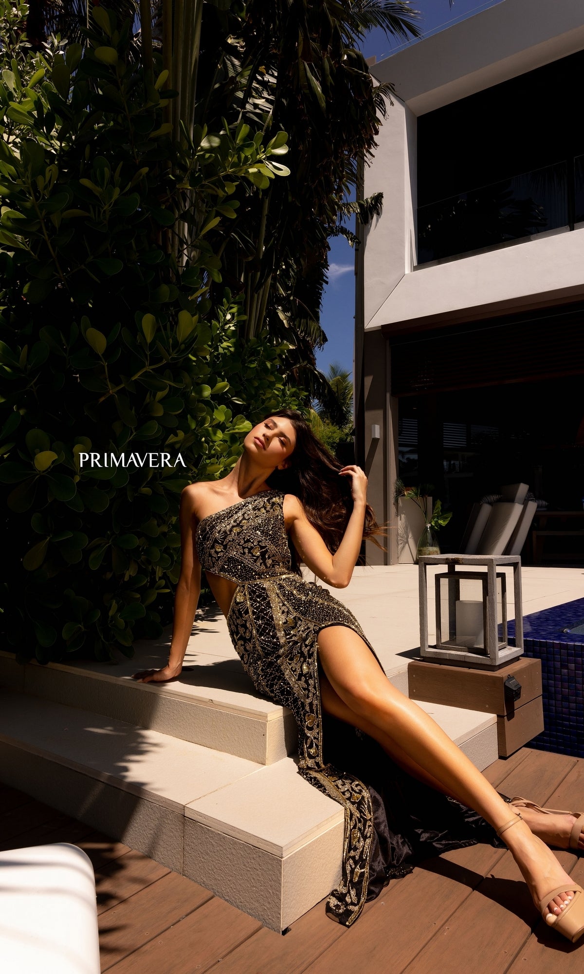 A woman in the One-Shoulder Primavera Beaded Long Prom Dress 4135 sits on outdoor steps by a modern house and pool, with