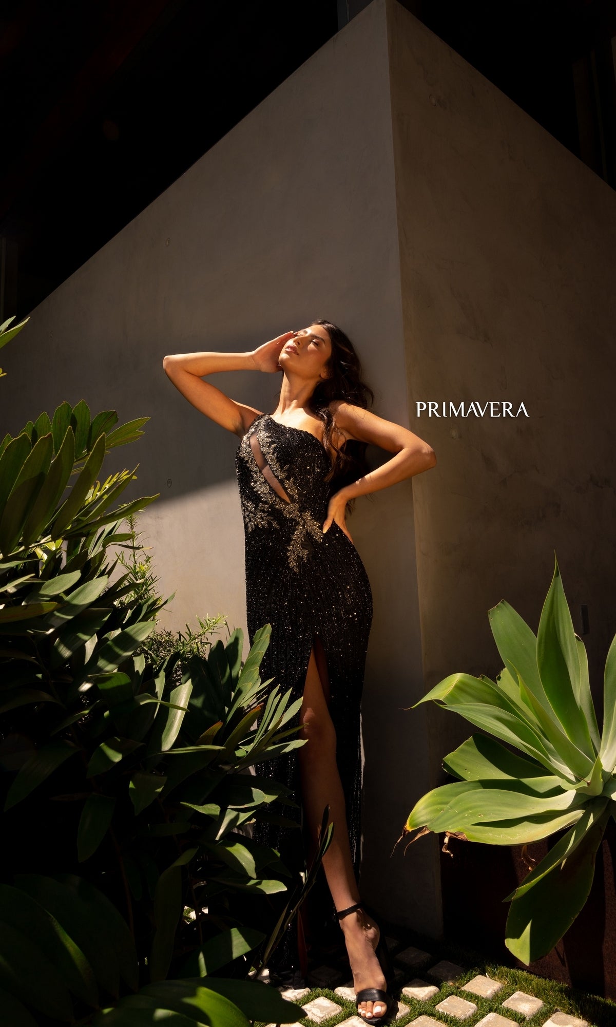 A woman poses outdoors by green plants against a concrete wall, wearing the Primavera One-Shoulder Beaded Long Prom Dress 4144—a black, sequined formal gown with a thigh-high slit that radiates elegance. The word
