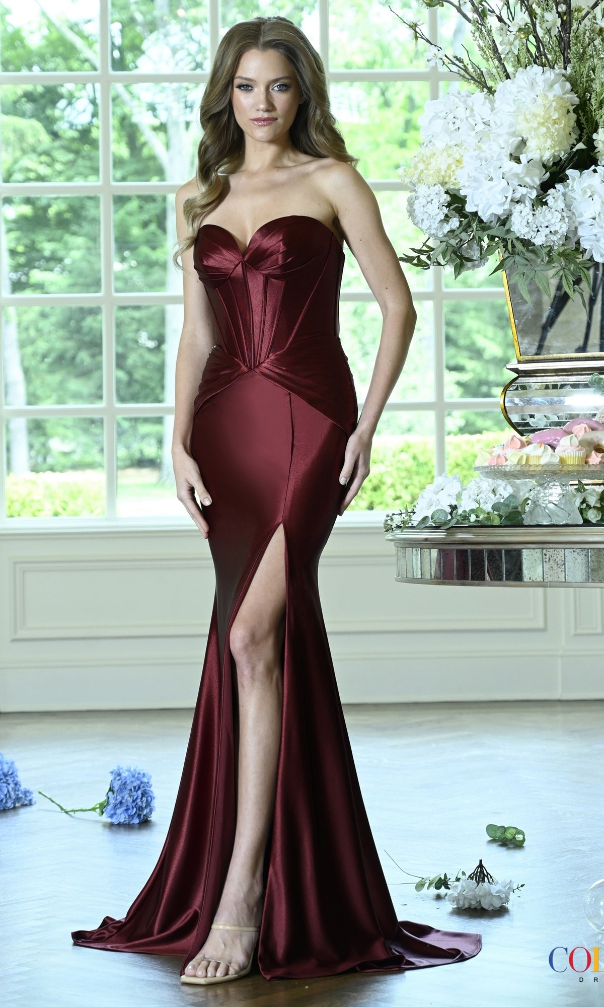 A woman wears a wine-colored Long Prom Dress: Colors Dress 4144, a floor-length strapless corset gown with a high thigh slit.