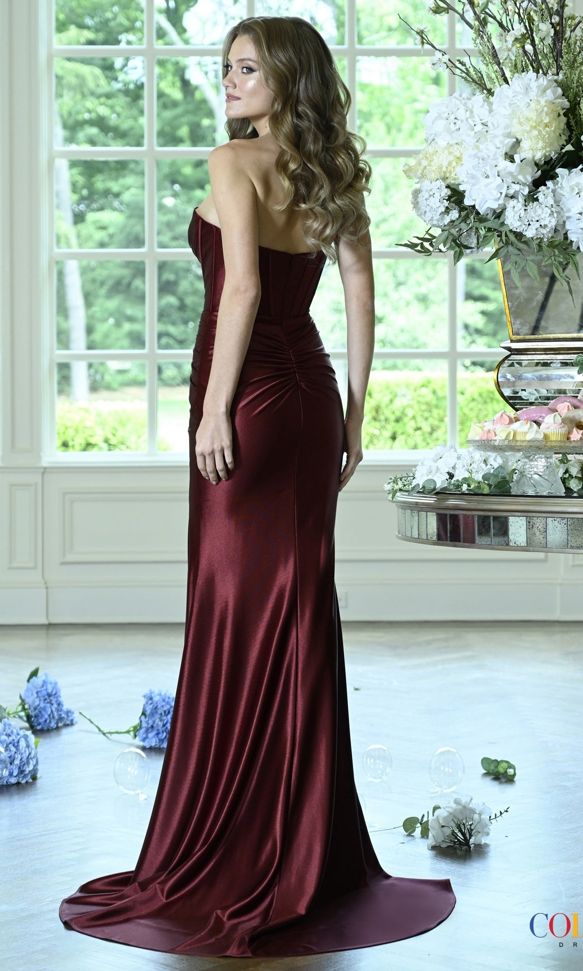 A woman wearing a floor-length, strapless burgundy satin mermaid Colors Dress 4144 prom dress with a corset-style bodice and a slight train.