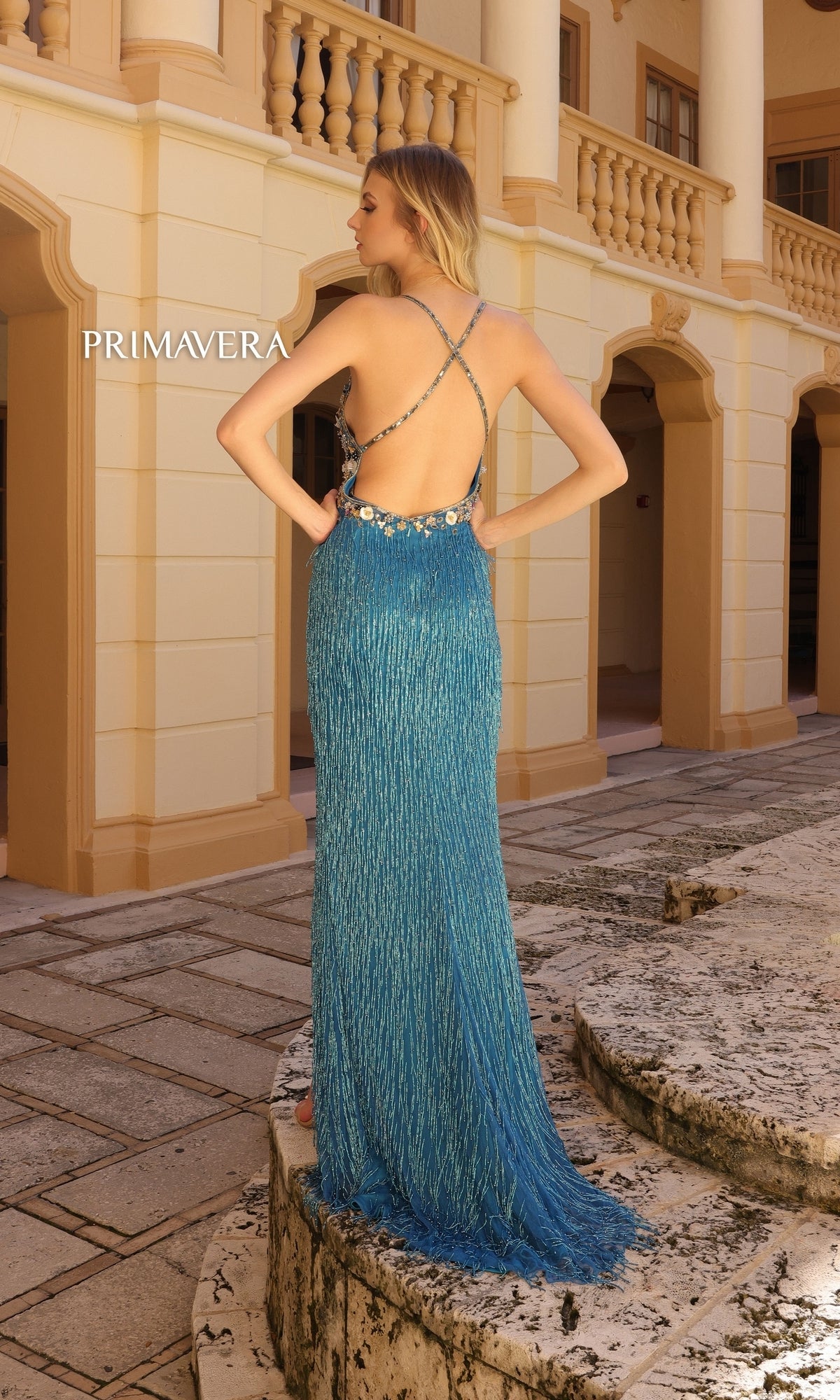 A woman in the Primavera Beaded-Fringe Long Prom Dress 4145, featuring a blue floor-length skirt, floral bodice, and beaded fringe, stands on a stone walkway before a beige columned building.