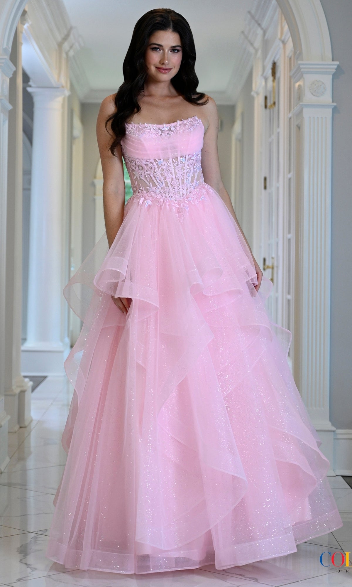 A model wears a pink Long Prom Dress: Colors Dress 4151, featuring a strapless lace corset bodice and a layered, glittery tulle skirt.