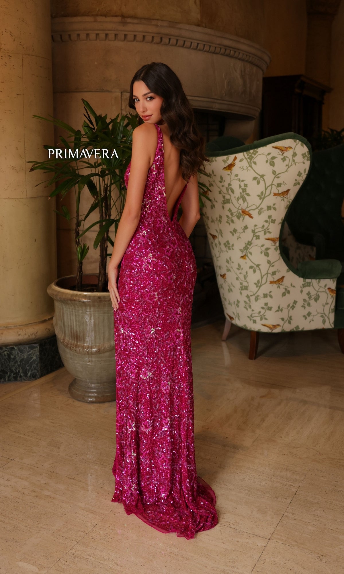 A woman poses indoors in the Primavera Star-Beaded Long V-Neck Prom Dress 4153, a deep pink sequined gown, looking over her shoulder in front of a large plant and an upholstered chair.