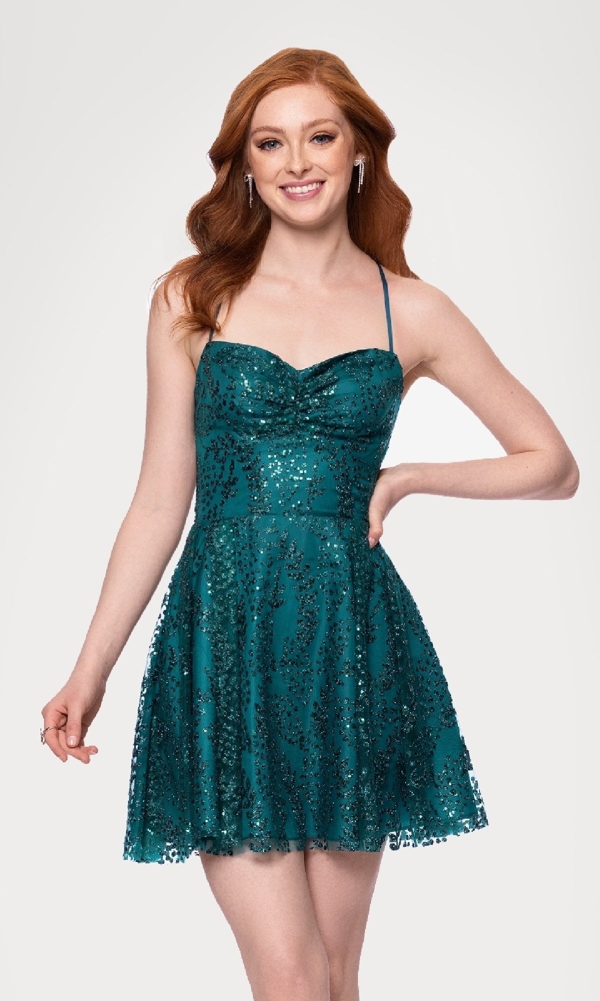 A woman with long red hair smiles in the Blondie Nites 4159BN Short Teal Glitter Hoco Dress, featuring a sleeveless design, against a plain white background.