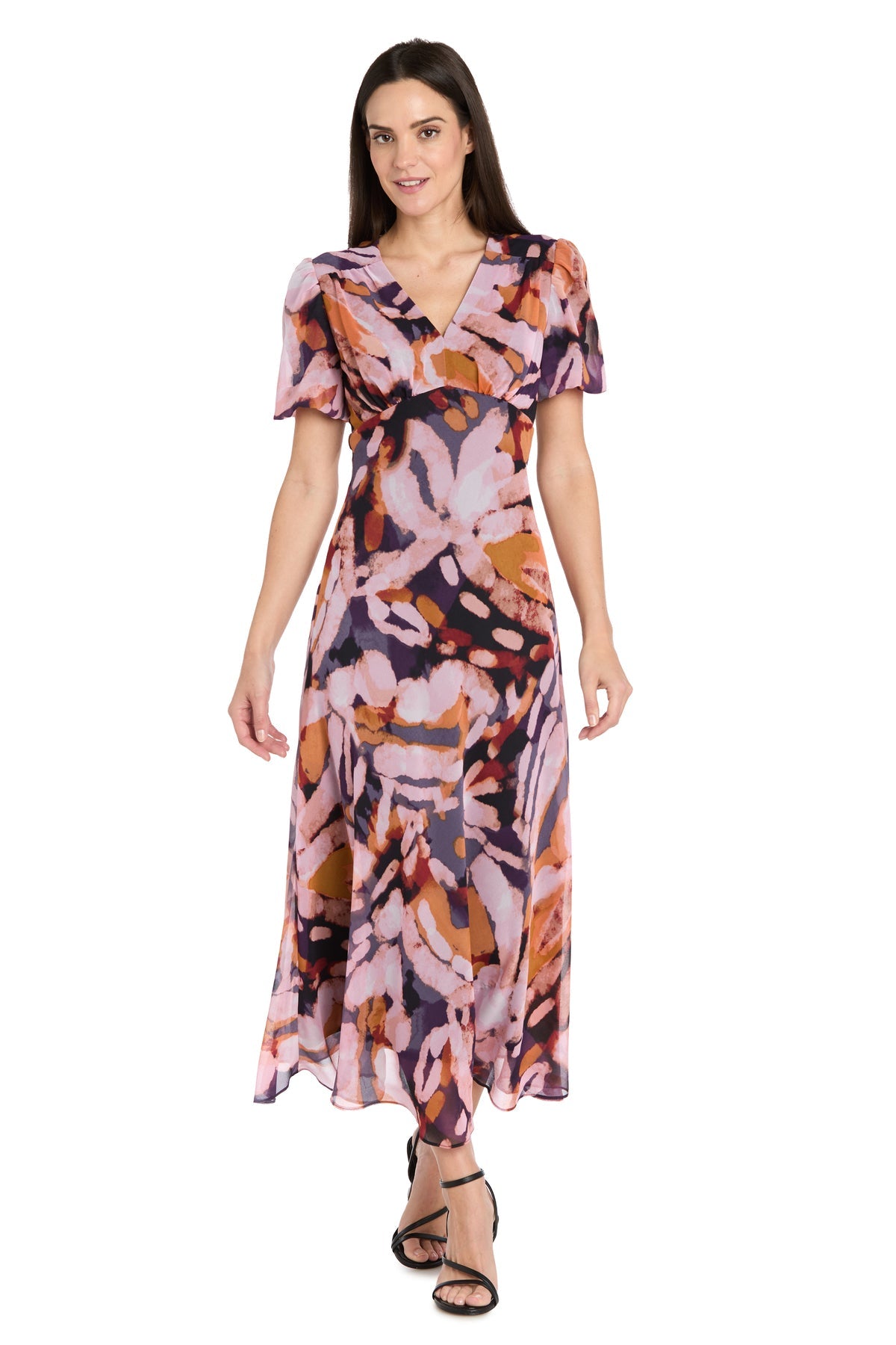 A woman wears the R&M Richards 4161 Short Sleeve Print Party Dress with a pink, purple, and orange abstract pattern, paired with black strappy sandals.