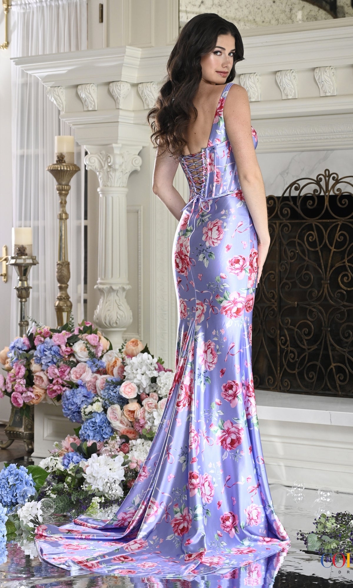 A woman wearing a Long Prom Dress: Colors Dress 4165, featuring a floor-length lavender gown with a pink rose print and a lace-up corset back.