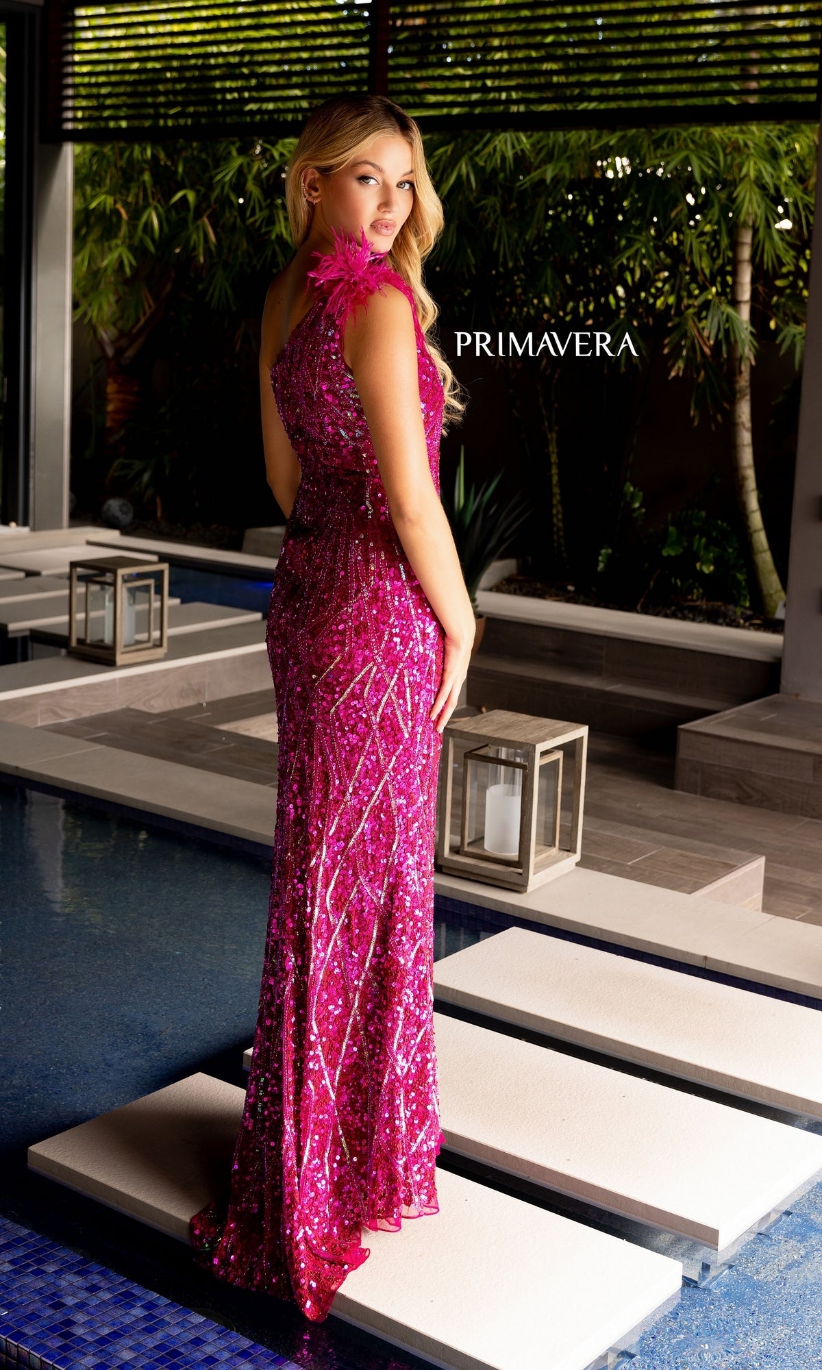 A woman in the Feather-Trim Primavera Long Sequin Prom Dress 4169 stands on a white walkway over a blue pool, surrounded by greenery and lanterns.