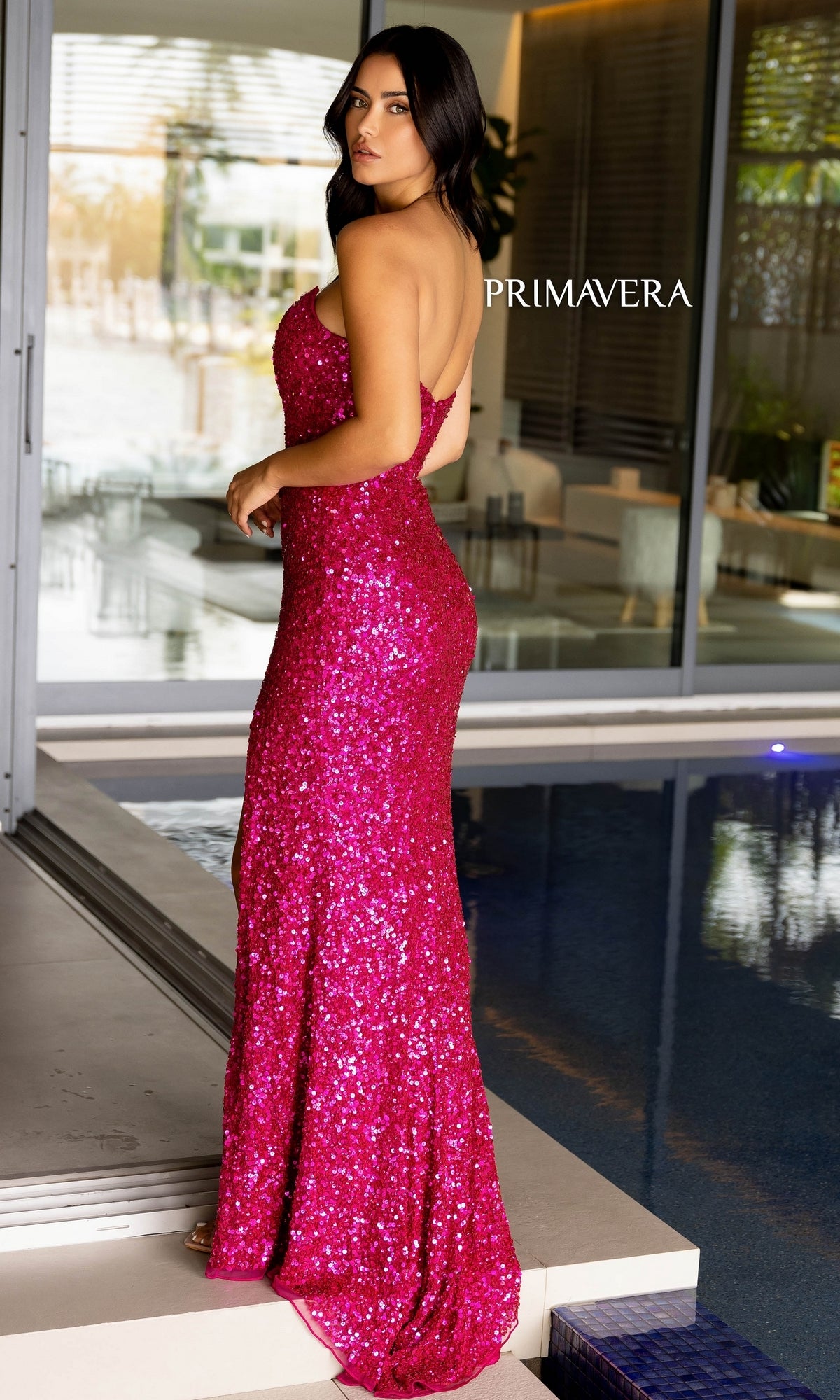 A woman wearing the Primavera Strapless Long Sequin Prom Dress 4192 stands by an indoor pool, gazing over her shoulder amid modern decor and large windows.
