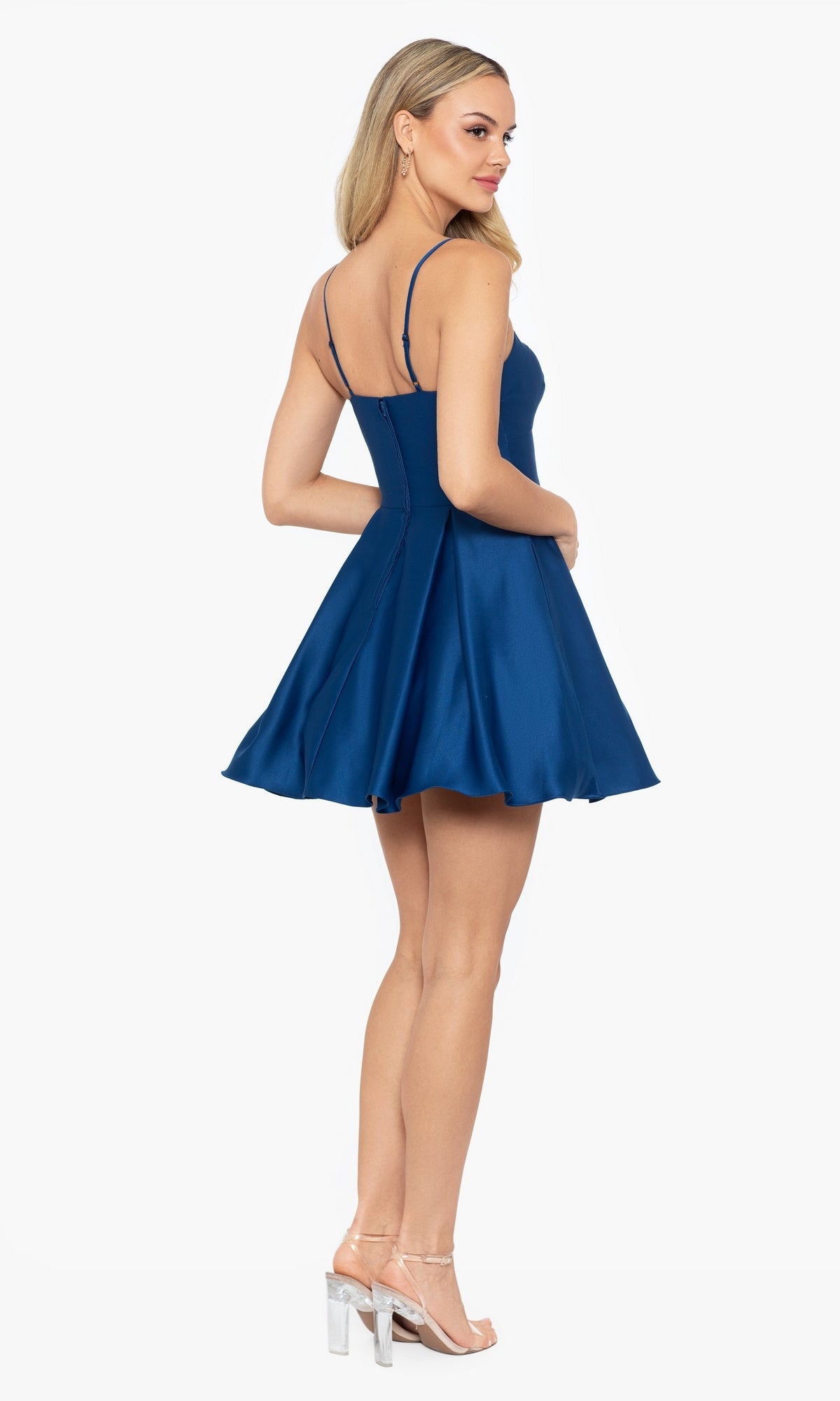 A woman stands looking over her shoulder in the Short Satin A-Line Homecoming Dress 4200BN, a sleeveless, flared mini dress, paired with clear high-heeled sandals.