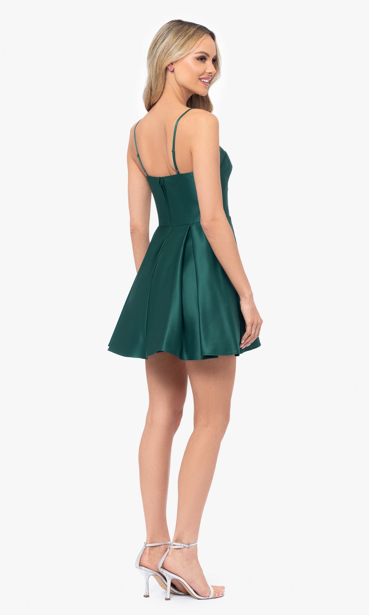 A woman models the Short Satin A-Line Homecoming Dress 4200BN, featuring thin straps and a pleated skirt, paired with silver high-heeled sandals against a plain white background.