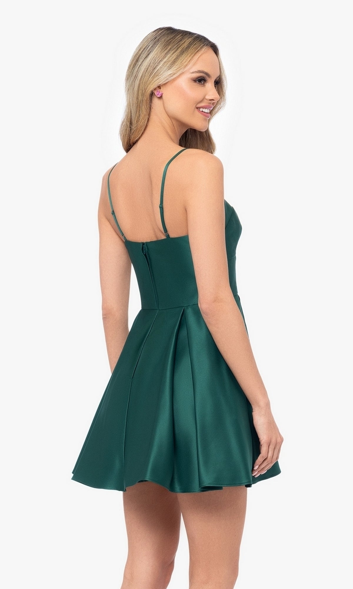 A woman with long blonde hair is wearing the Short Satin A-Line Homecoming Dress 4200BN, a sleeveless dark green dress with spaghetti straps, standing and looking over her shoulder.