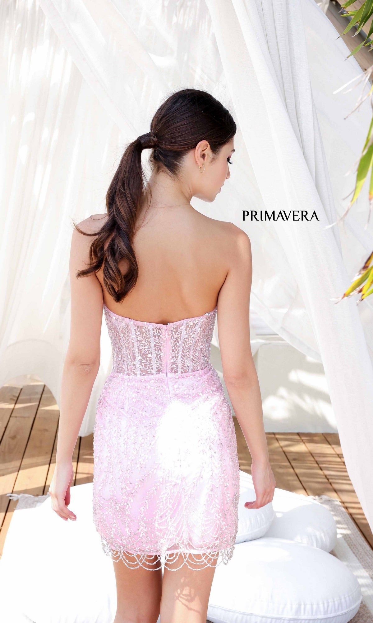 A woman with brown hair in a ponytail wears the Short Homecoming Dress 4213 by Primavera in light pink lace, standing with her back to the camera near white curtains and cushions.