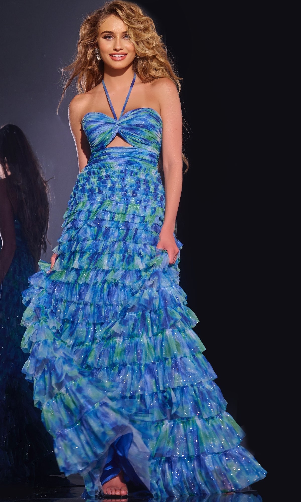 A woman stands barefoot against a dark background, smiling with loose wavy hair, wearing the Jovani 42182 Long Prom Dress in blue and green with tiered ruffles.
