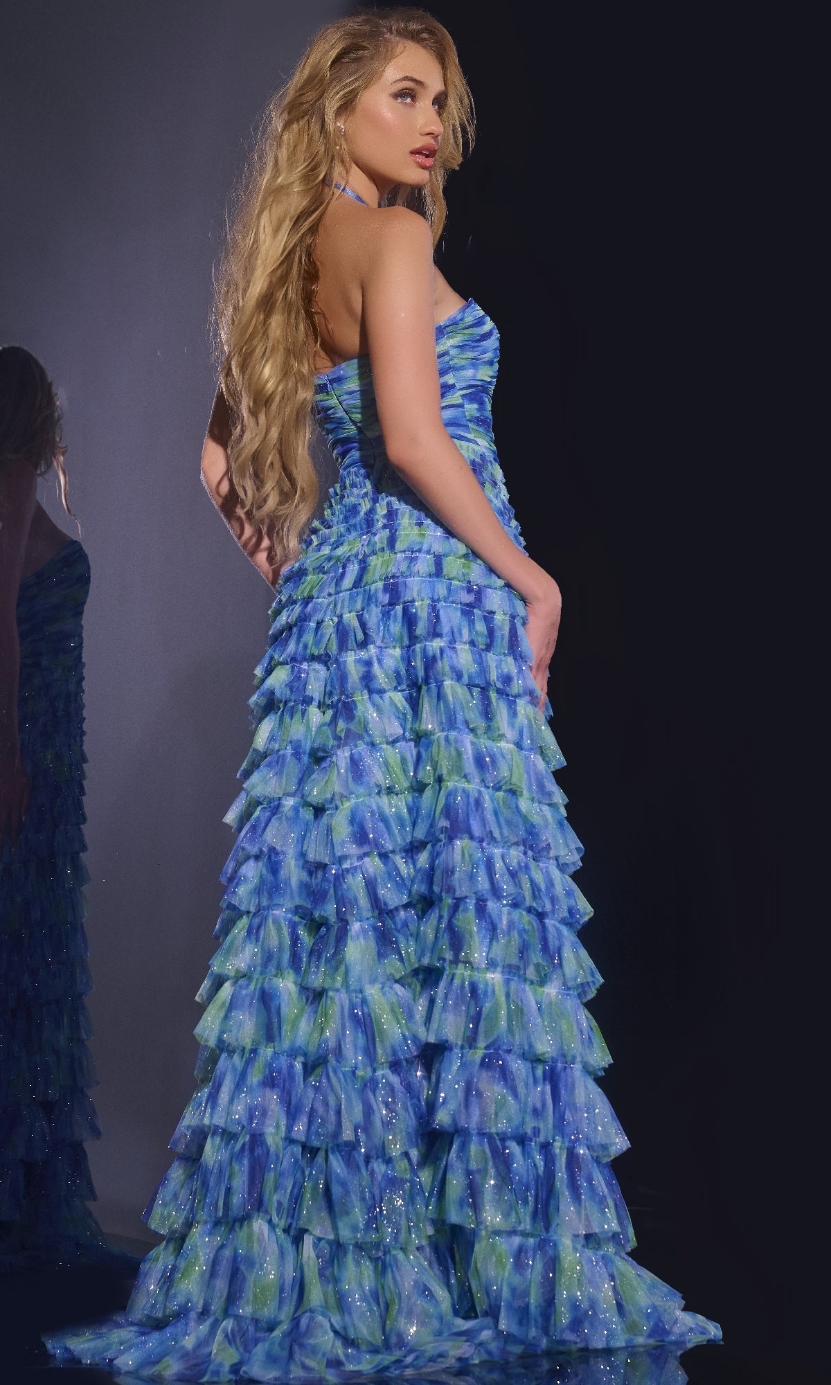 A woman stands sideways against a dark background wearing the Jovani 42182 long prom dress, a strapless, floor-length gown with blue and green watercolor patterns and an elegant tiered ruffle design.